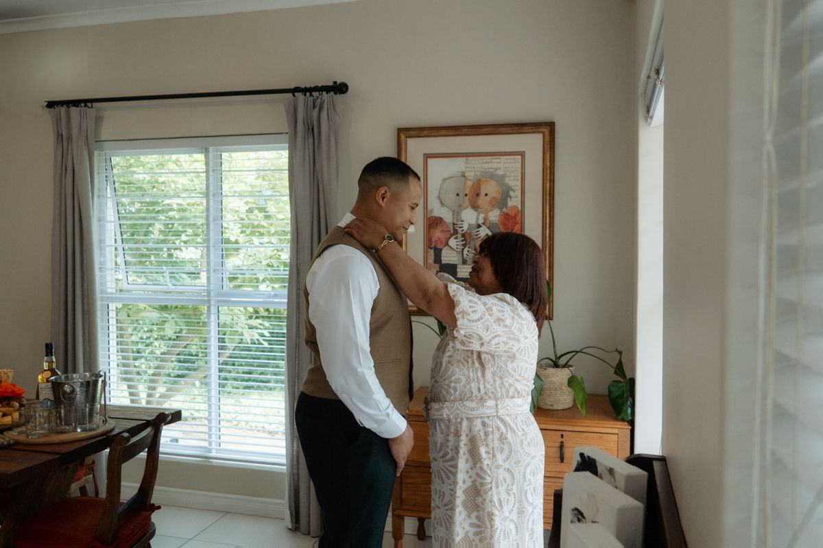 Woman helping groom adjust his boutonniere near a bright window before the wedding ceremony.