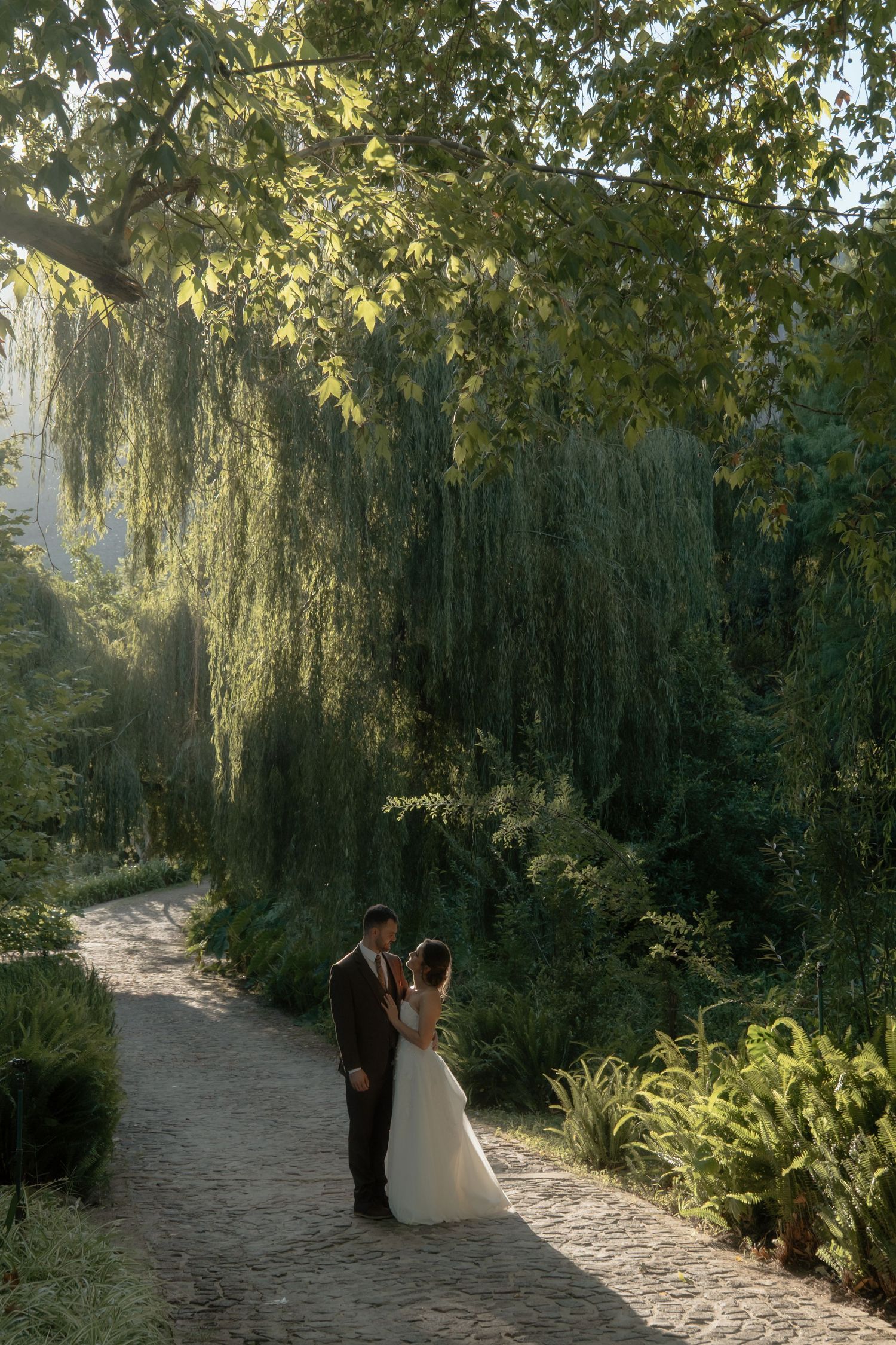 Bride and groom share a romantic moment on a cobblestone path beneath a weeping willow tree.