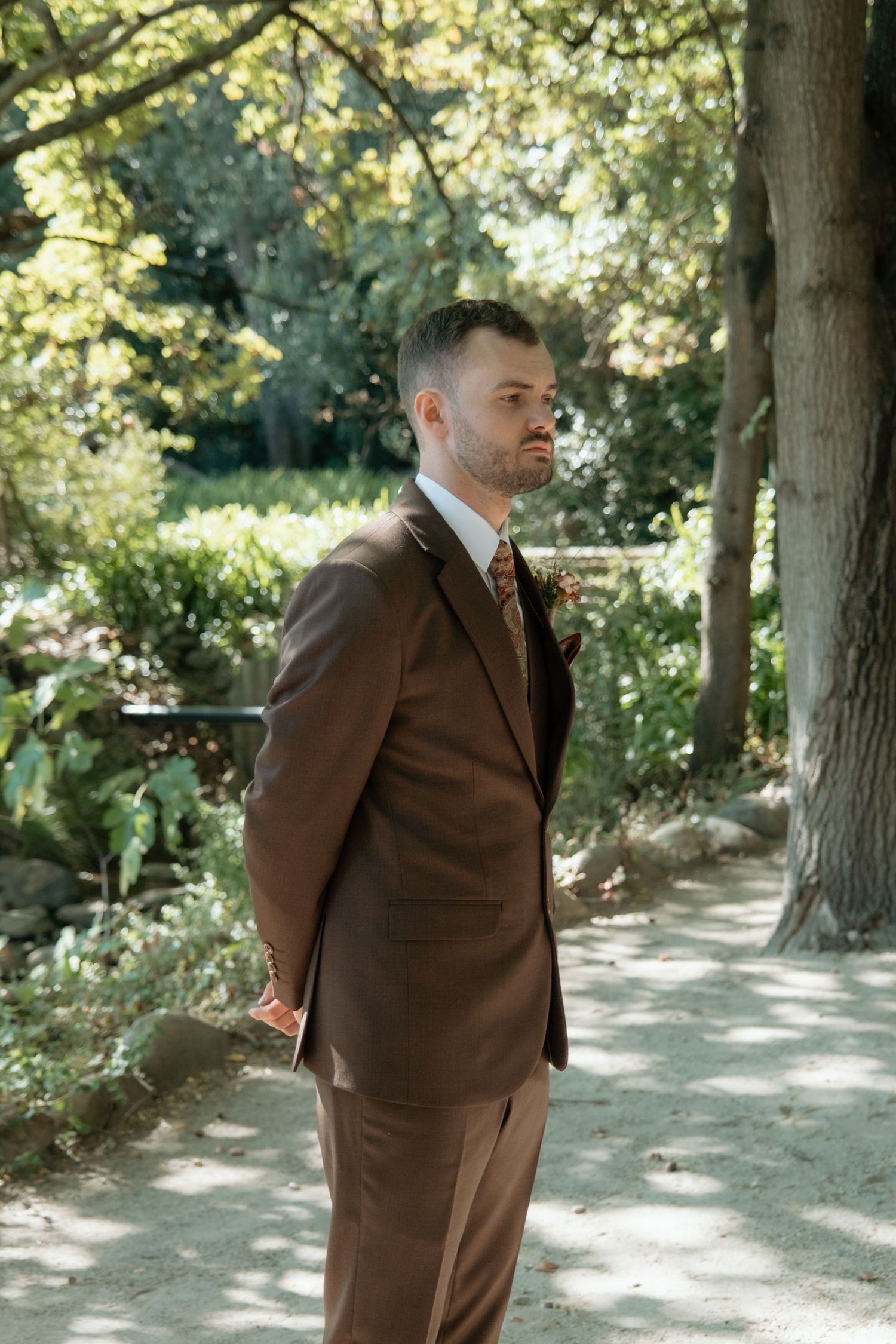 Groom in a brown suit standing confidently on a garden path surrounded by lush green trees.