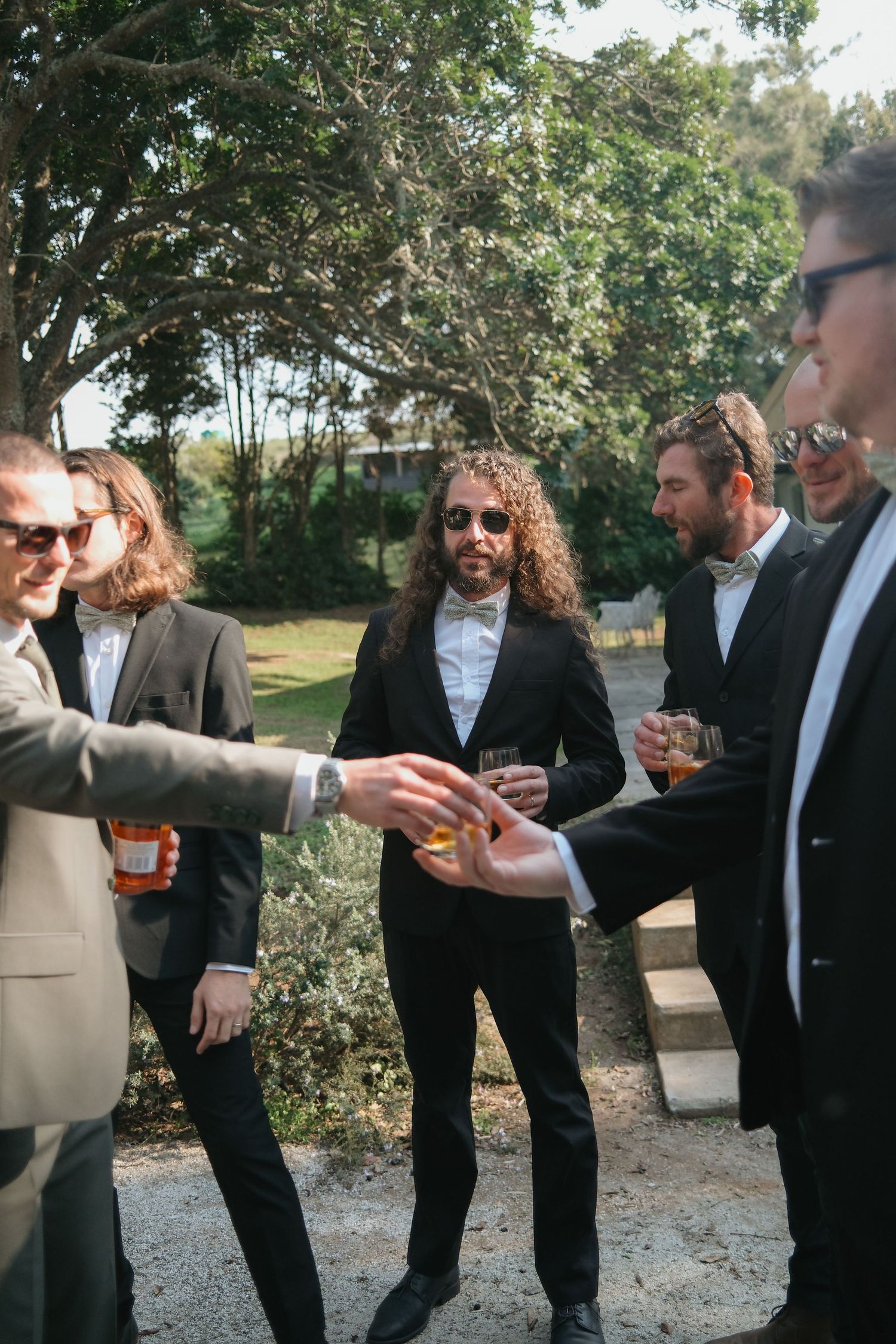 Groomsmen in suits toasting with drinks outdoors at a wedding celebration.