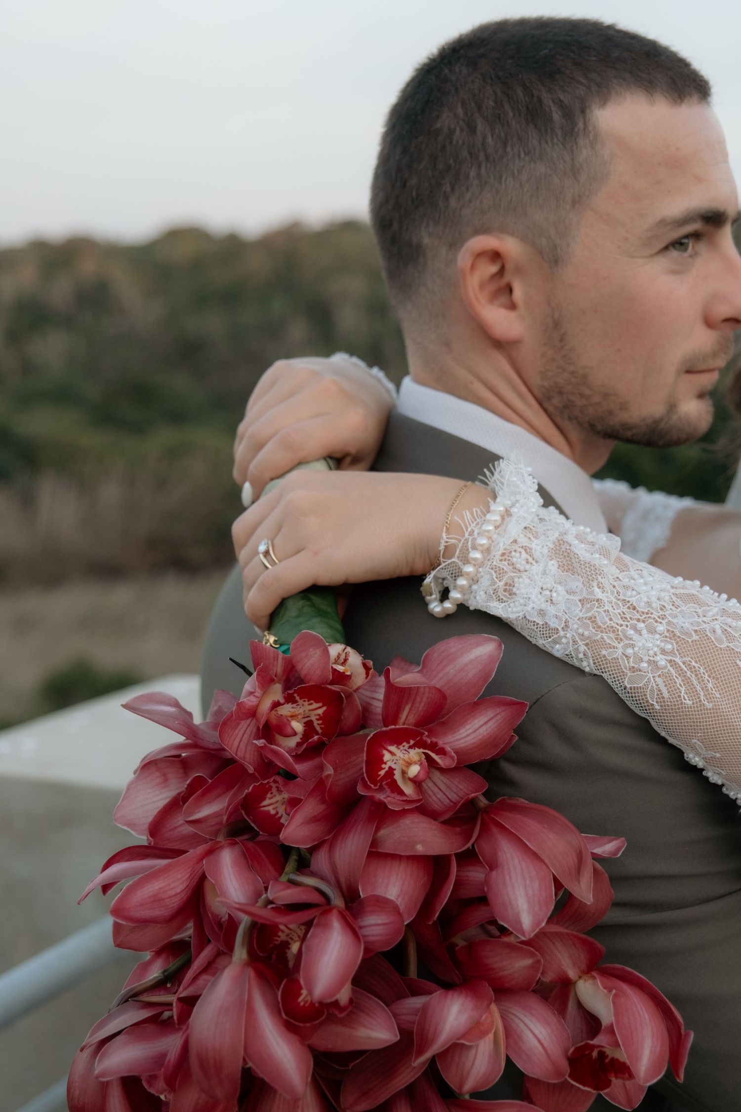 Groom in gray suit embraced by bride holding deep red orchid bouquet at outdoor wedding.