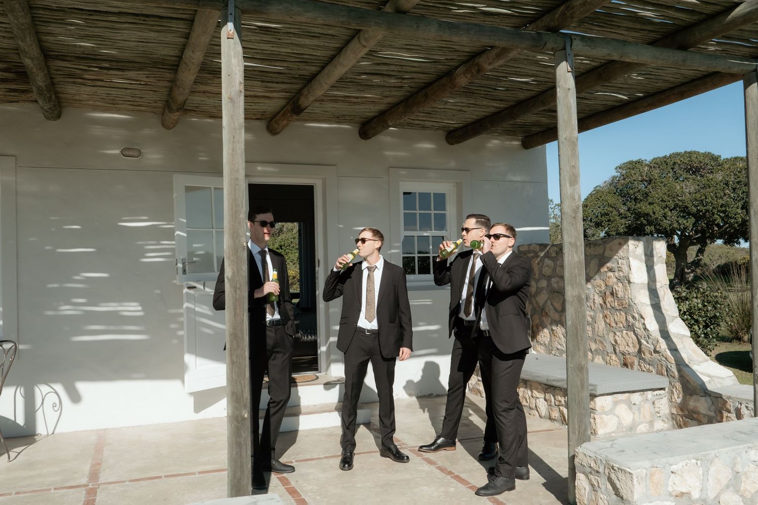 Four groomsmen in black suits toasting drinks outside a white Mediterranean villa on a sunny day.