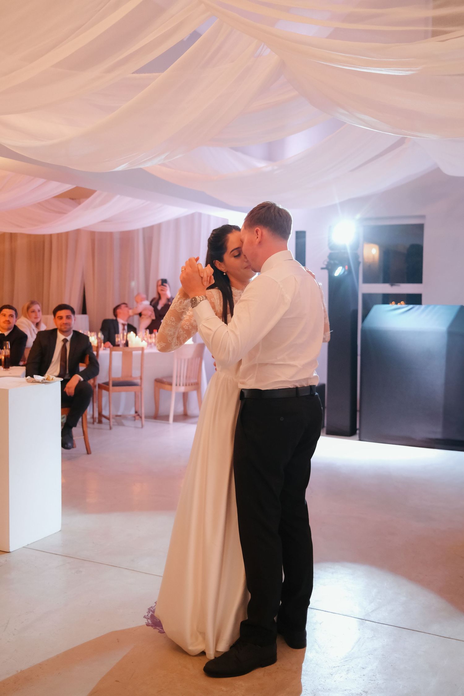 Bride and groom share their first dance in elegant draped wedding venue with guests watching.