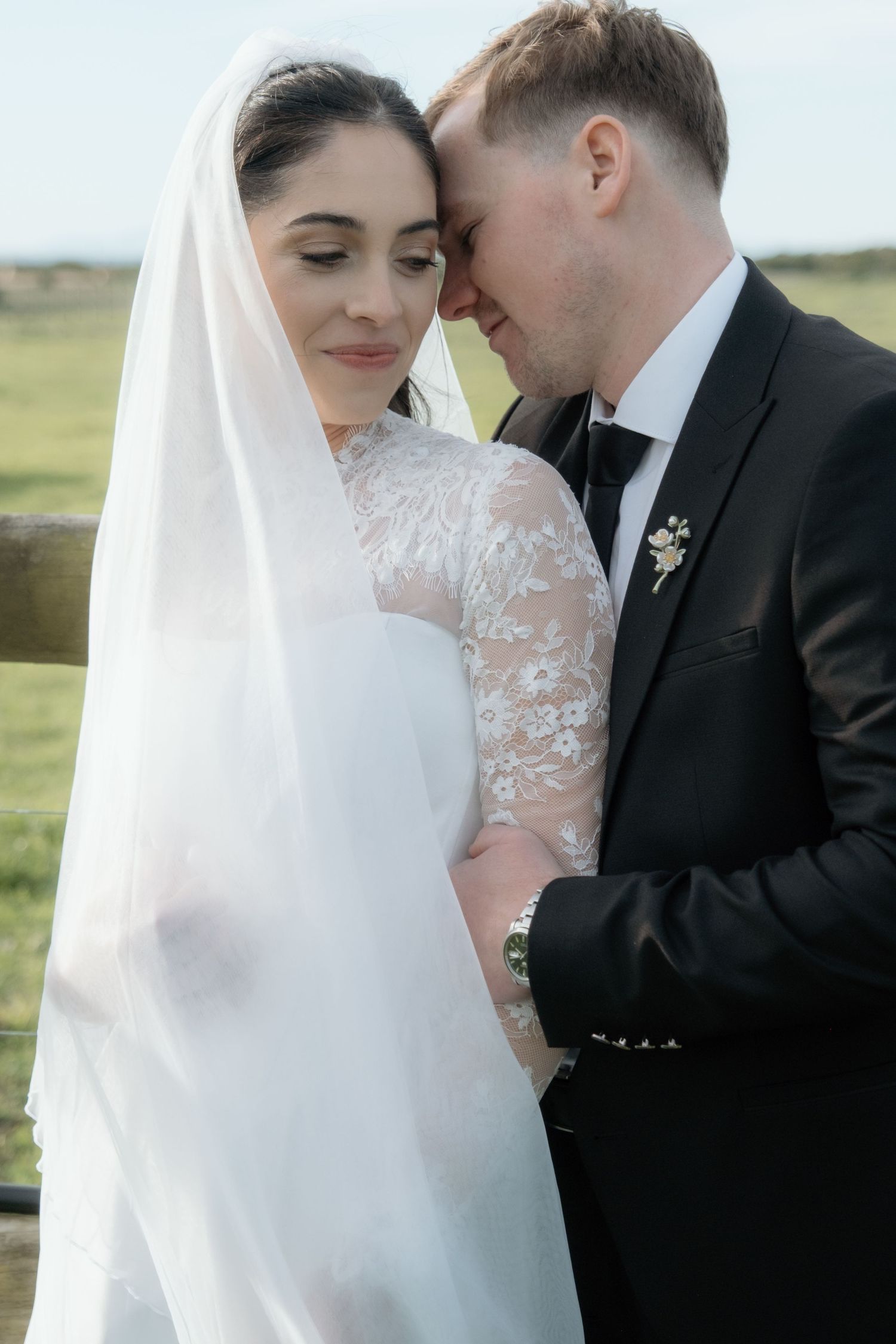 Bride and groom share an intimate moment, touching foreheads, bride wearing lace gown and veil outdoors.