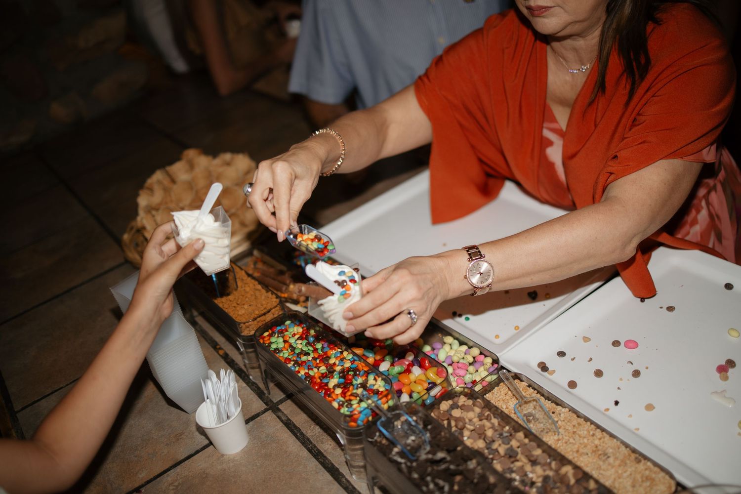 Woman in orange top serving candy toppings at a dessert bar, with colorful sprinkles and chocolates on display.