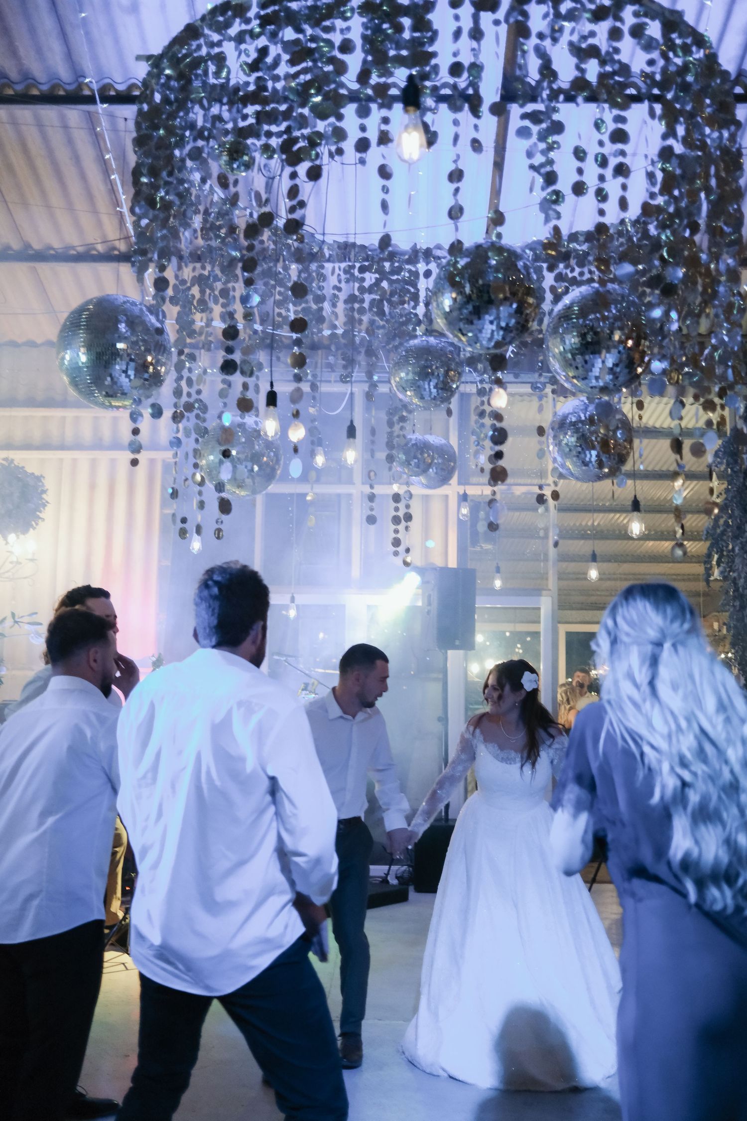 Bride and groom dance with guests under sparkling disco balls and hanging decorations at a wedding reception.