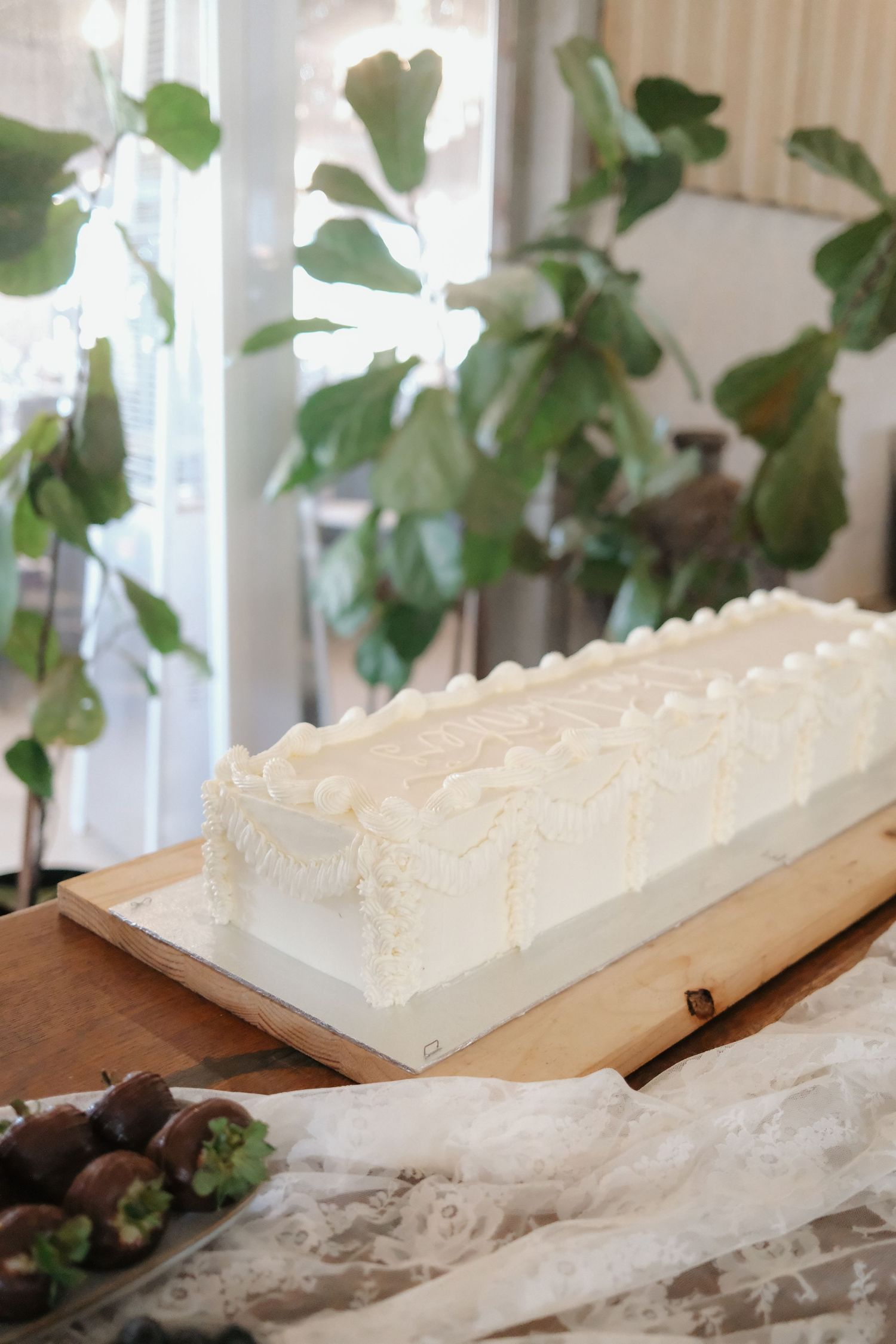 Elegant rectangular white frosted wedding cake on wooden board surrounded by green foliage.