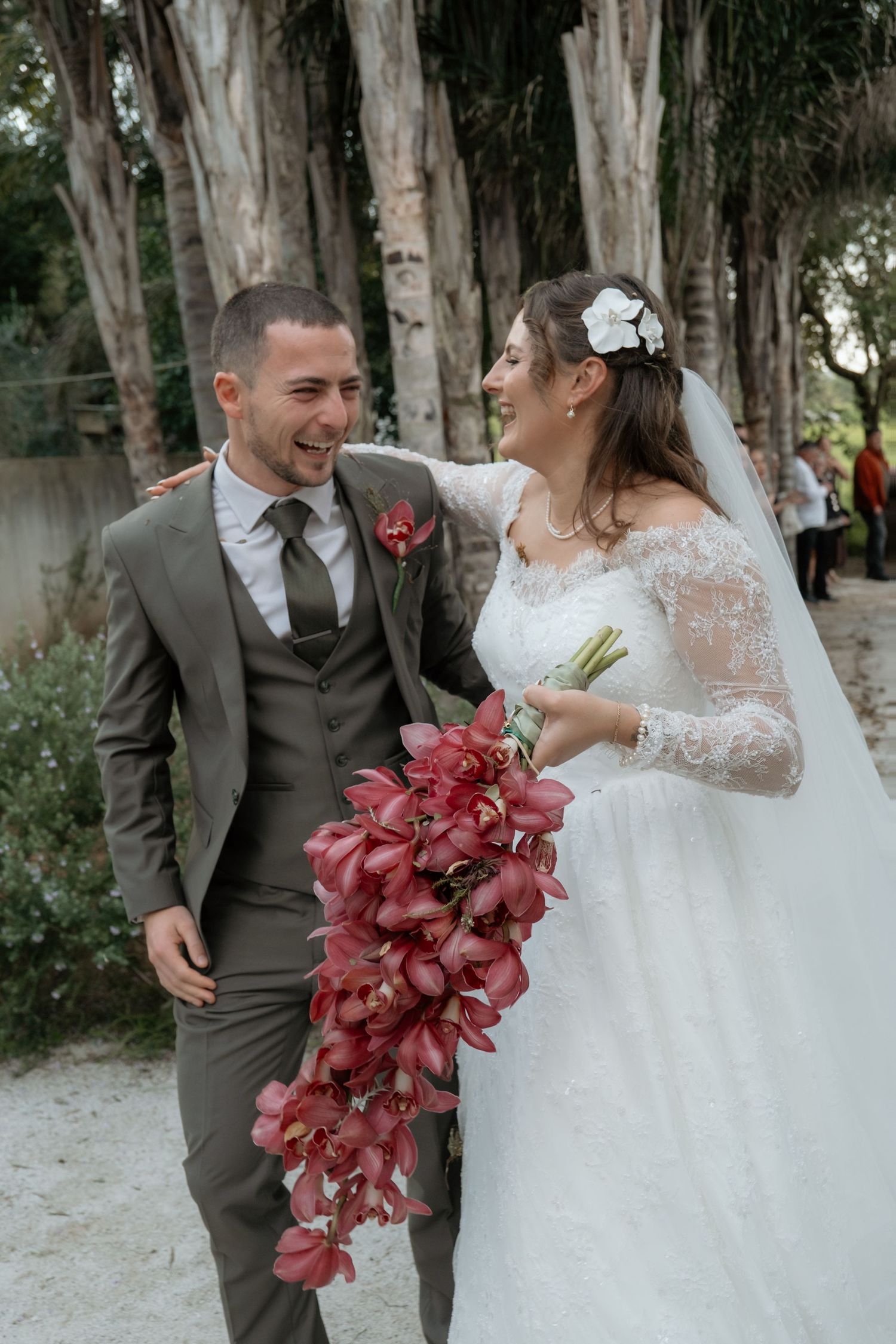 Laughing bride and groom walk together after ceremony, bride holding cascading red orchid bouquet.