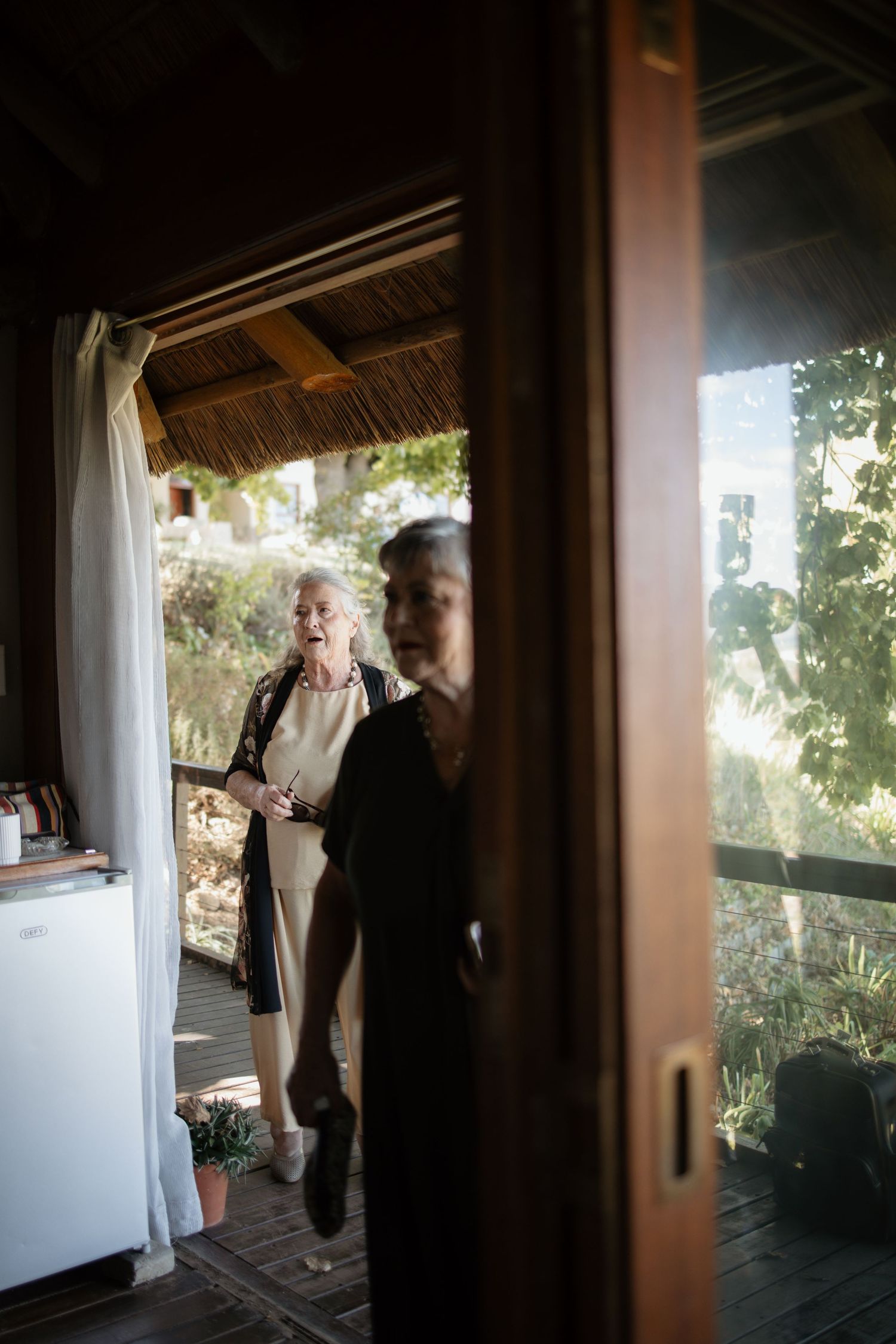 Elderly woman in black jacket standing on wooden porch of rustic thatched cottage surrounded by lush greenery.