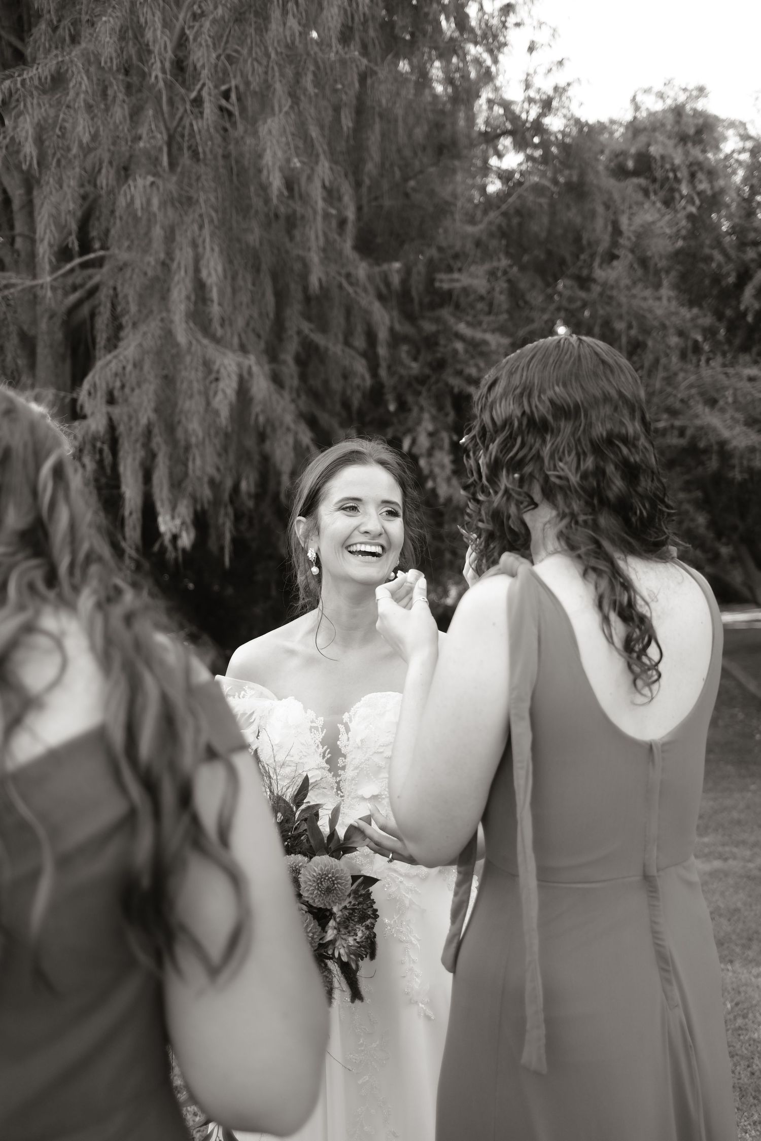 Laughing bride in lace wedding dress shares joyful moment with bridesmaids in black and white.