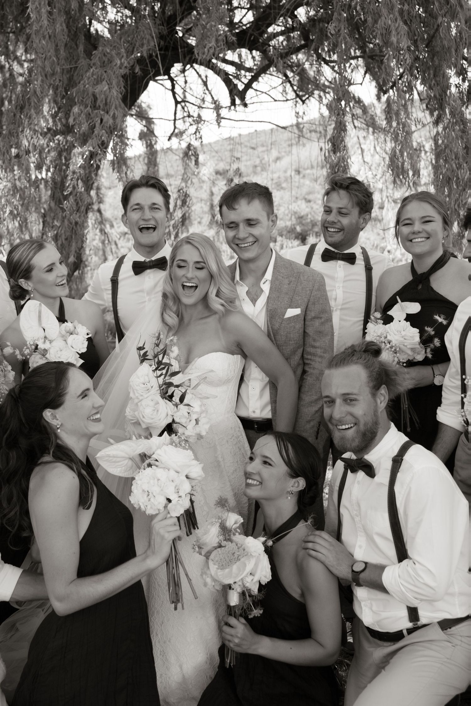 A wedding party in formal attire poses together under a willow tree, creating a joyful black and white portrait.