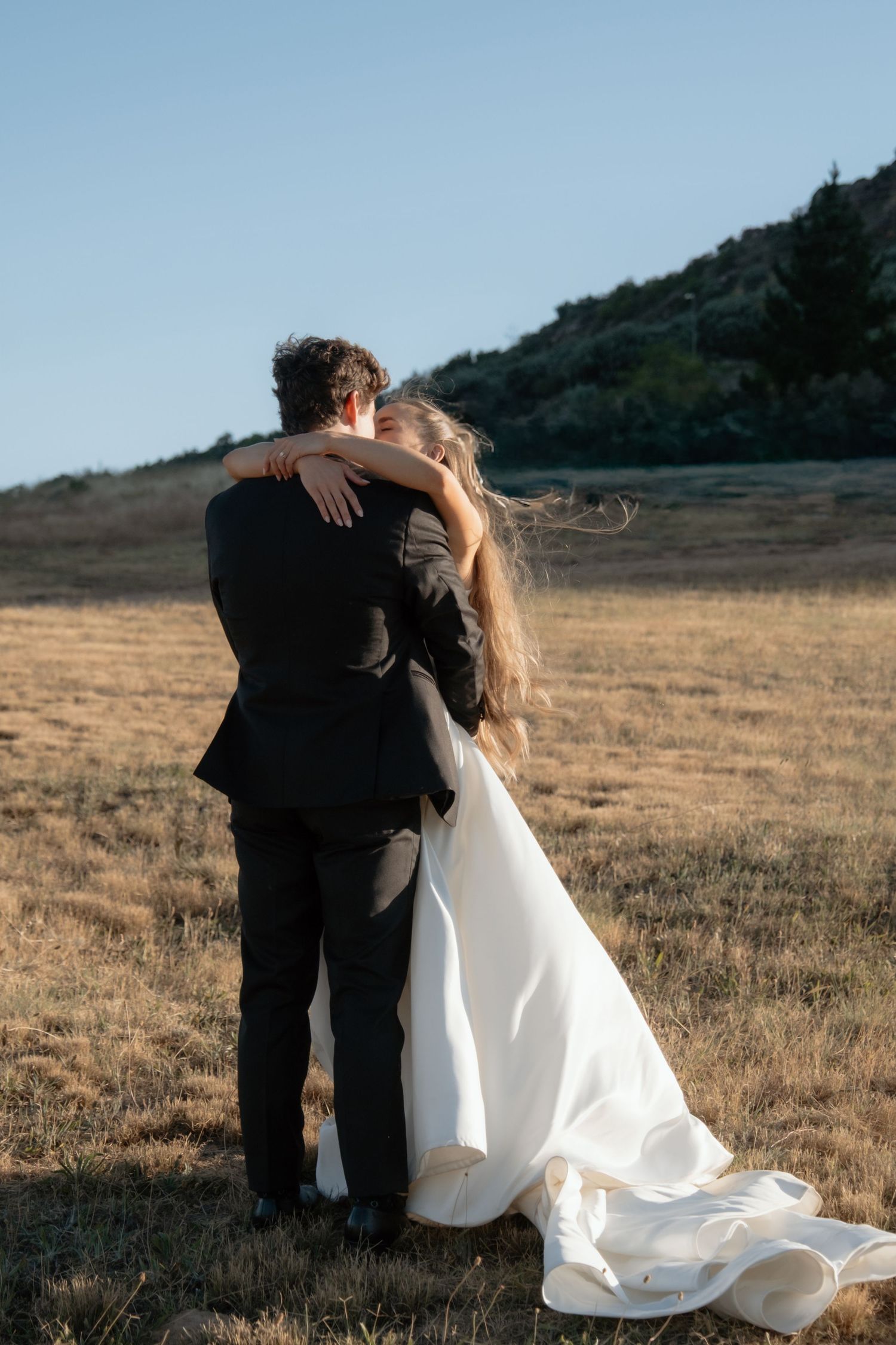 A bride and groom embrace in a golden field with rolling hills, sharing a romantic moment in their wedding attire.