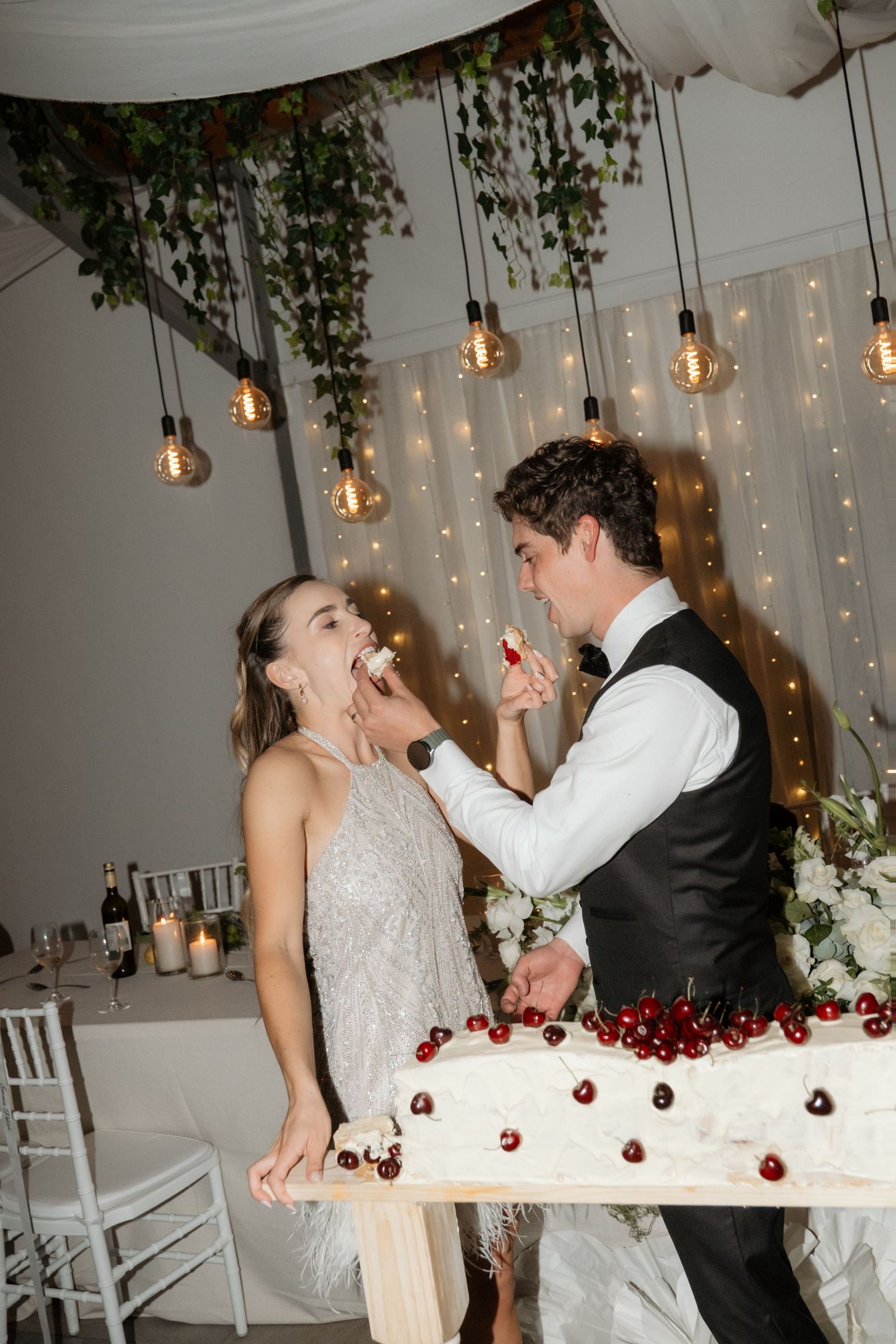 Groom in tuxedo feeding bride a cherry from elegant white wedding cake at reception celebration.