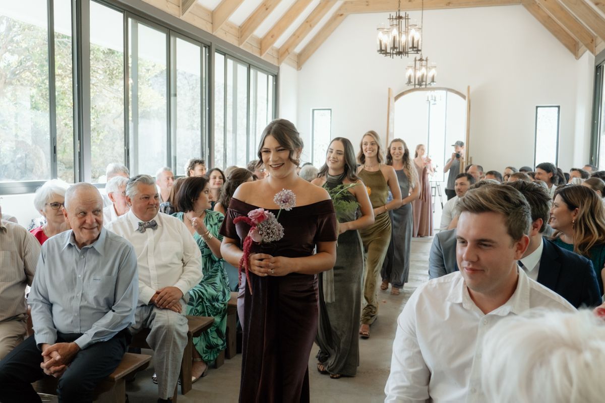 Bridesmaids in dark velvet gowns walk down the aisle at an elegant indoor wedding ceremony with guests seated.