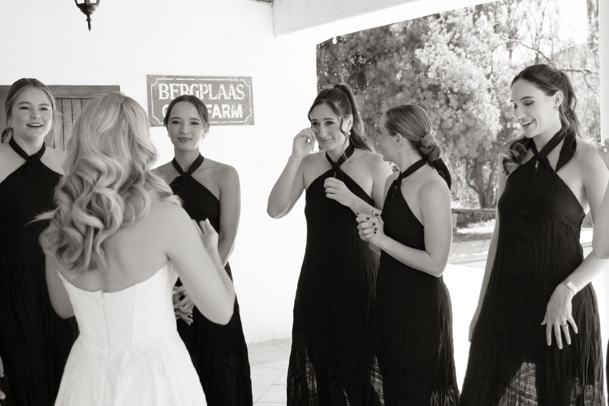 Black and white photo of bride in wedding dress revealing gown to emotional bridesmaids in black dresses outdoors.