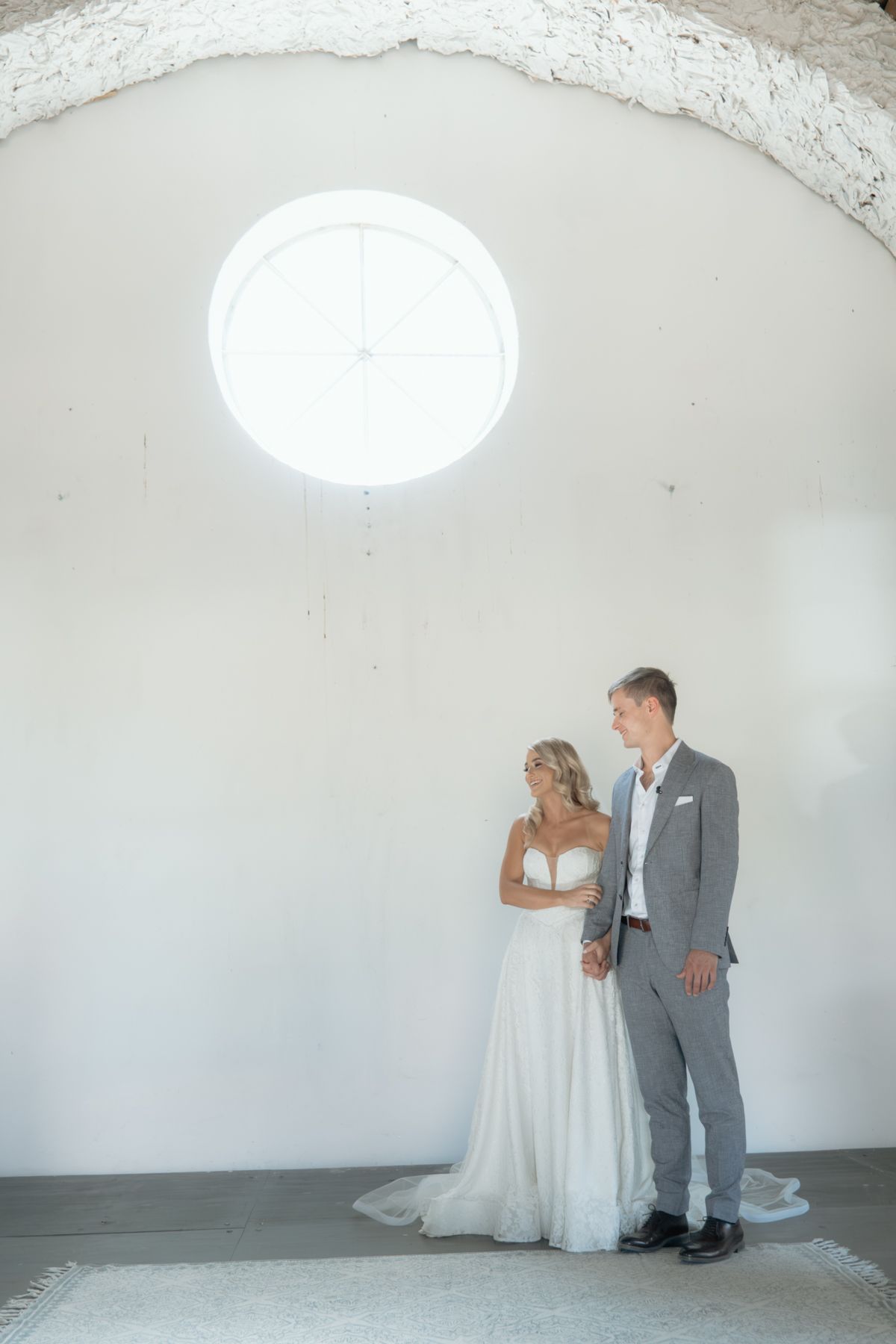 Bride and groom standing together under a white arched ceiling with a round window in a minimalist chapel.