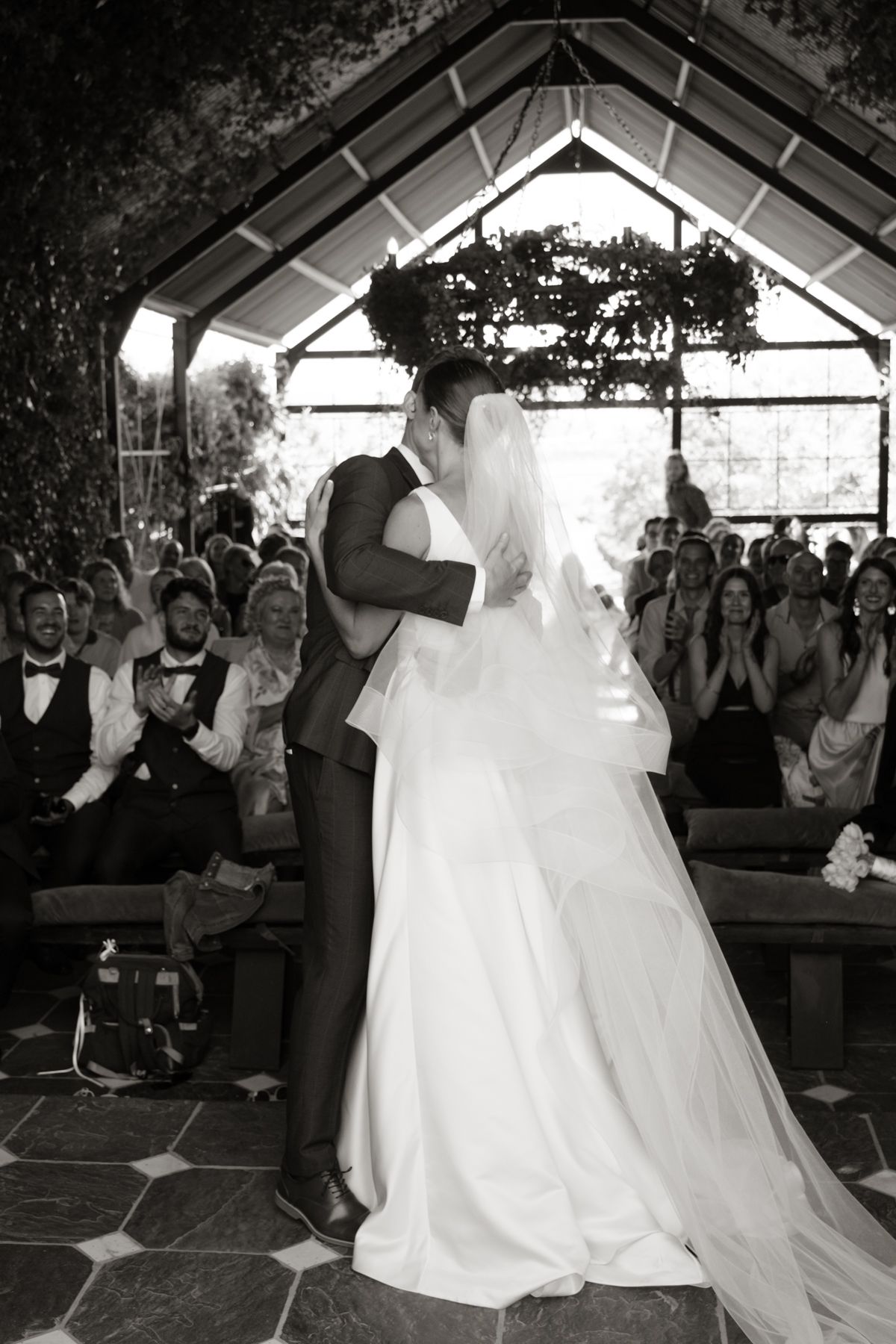 Black and white photo of bride and groom sharing first kiss at the altar in a glass chapel.