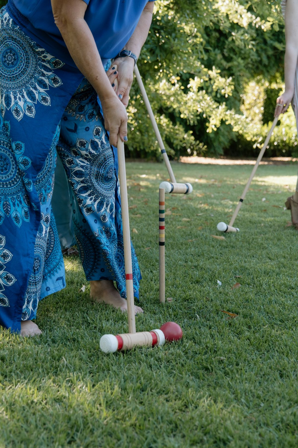 A person in blue patterned pants plays croquet on a lush green lawn in a sunny garden setting.