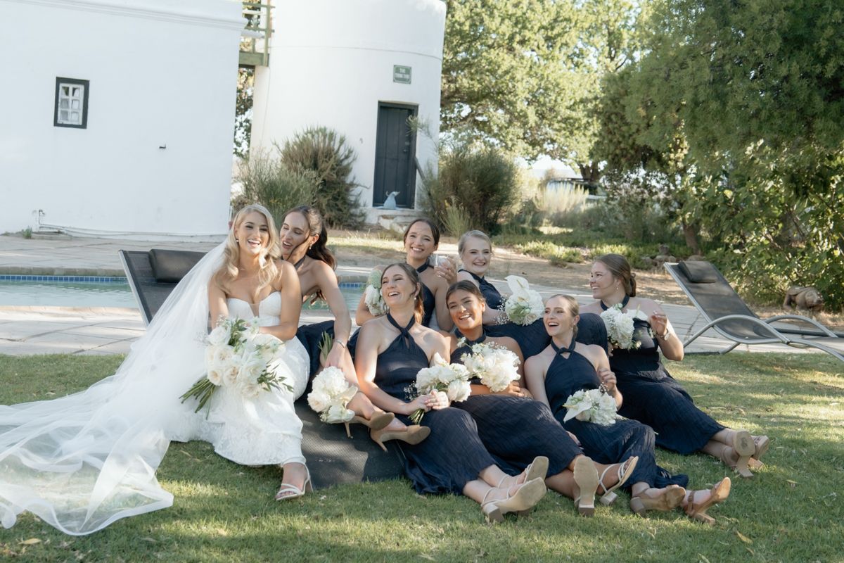 Bride and bridesmaids in navy dresses laughing together on grass holding white bouquets at outdoor wedding venue.