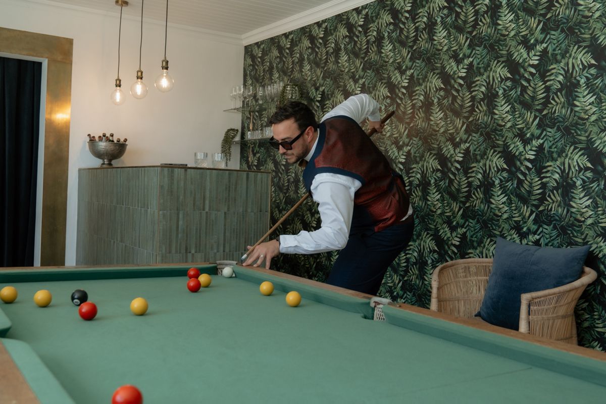 Man in vest and sunglasses playing pool on green billiard table in stylish room with tropical wallpaper.