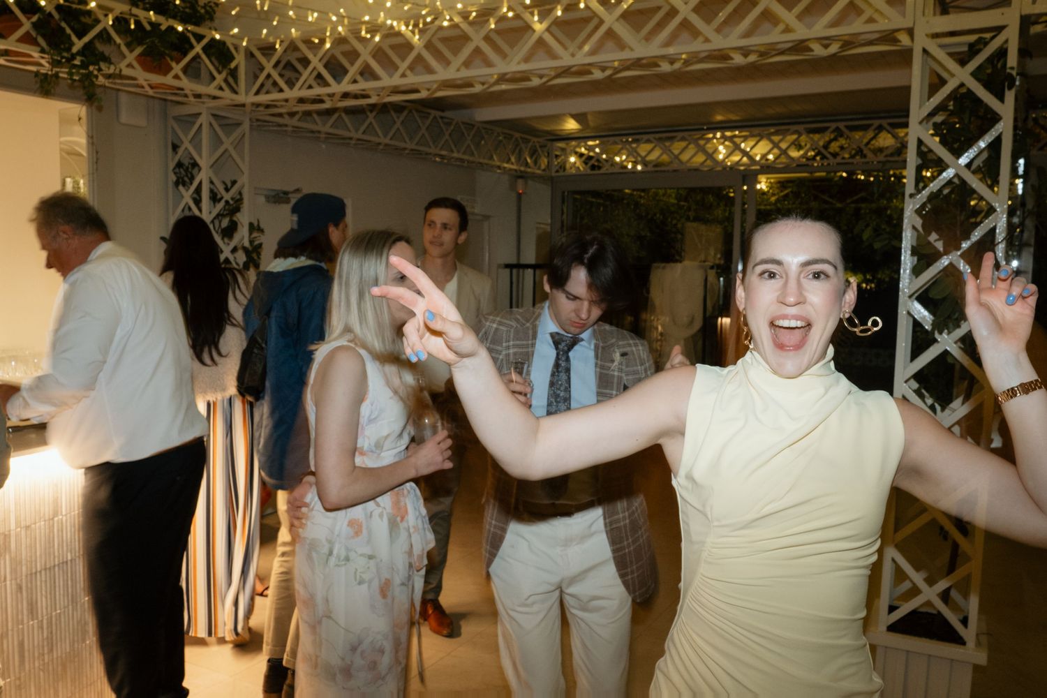 Excited wedding guest dances with arms raised at a festive indoor wedding reception with fairy lights.
