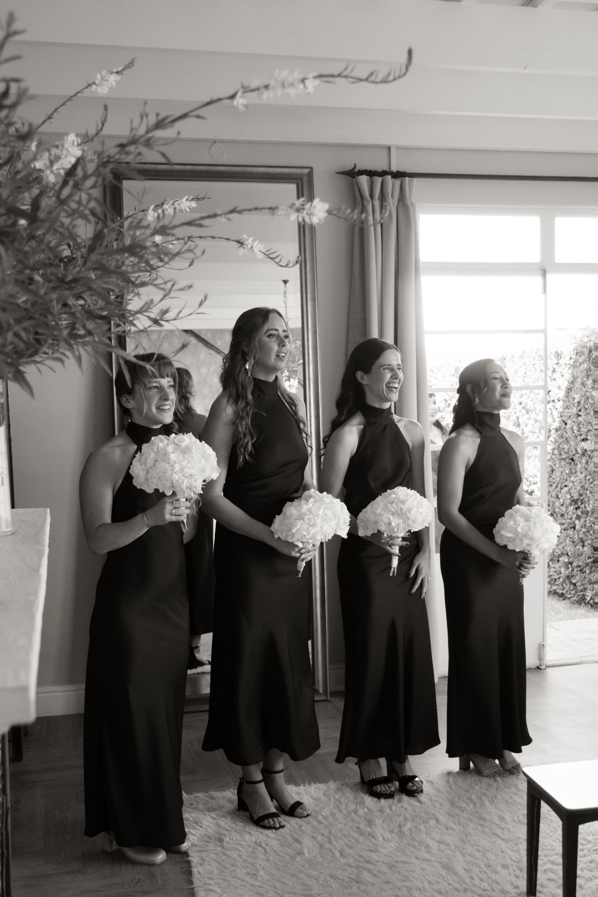 Four bridesmaids in black halter gowns hold white hydrangea bouquets in a black and white wedding photo.