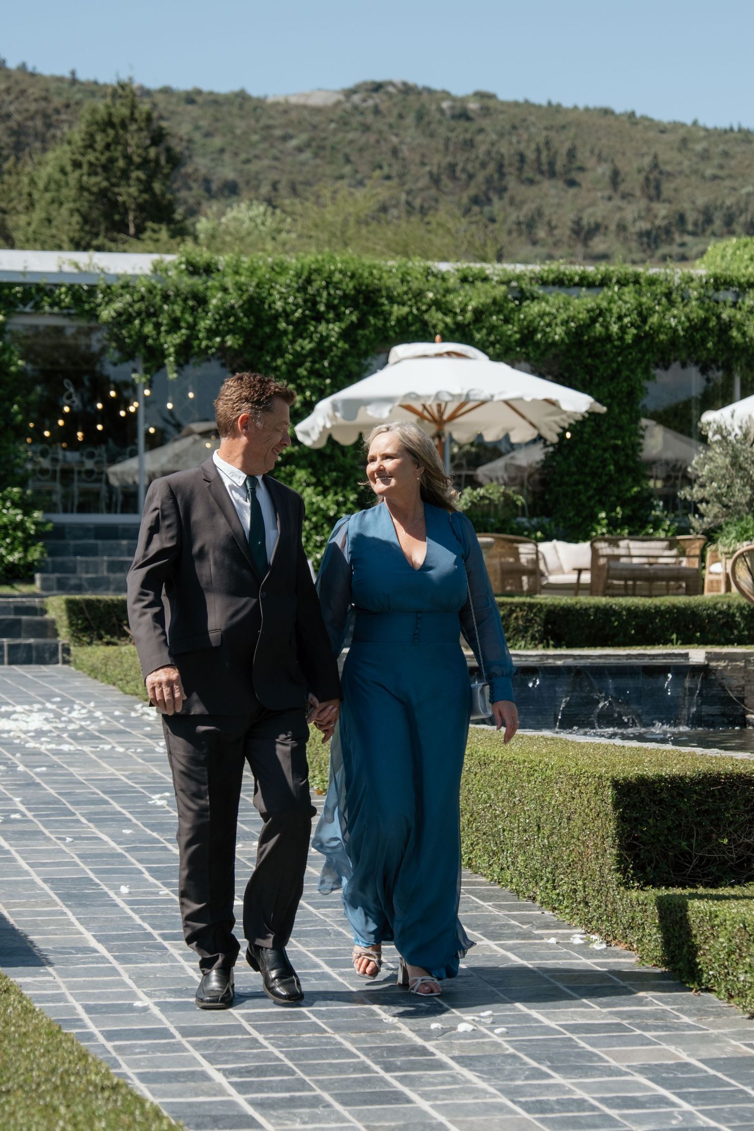 Couple walking hand in hand along a garden path, man in dark suit, woman in teal dress, lush greenery backdrop.