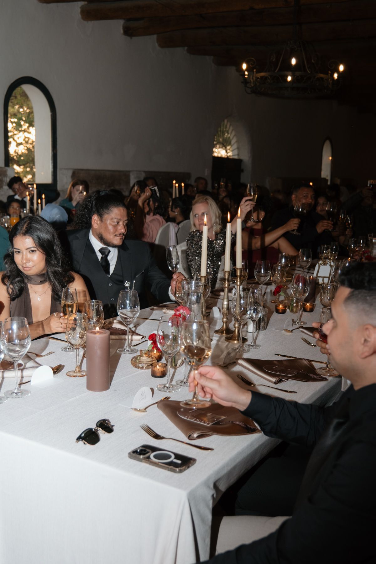 Wedding reception guests seated at elegantly decorated tables with candles and glassware in a rustic venue.