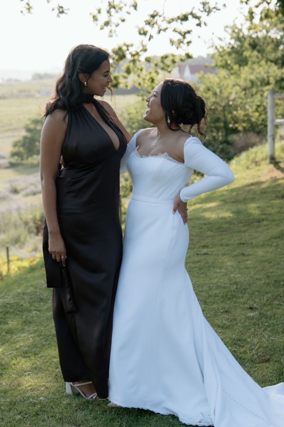 Bride in white mermaid gown poses with bridesmaid in black halter dress in garden setting.