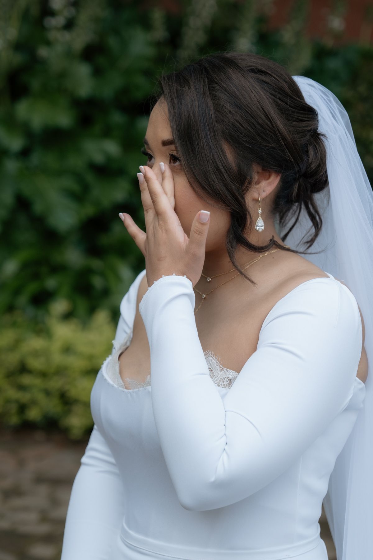 Emotional bride in white long-sleeve gown and veil covers her mouth with hand outdoors on wedding day.