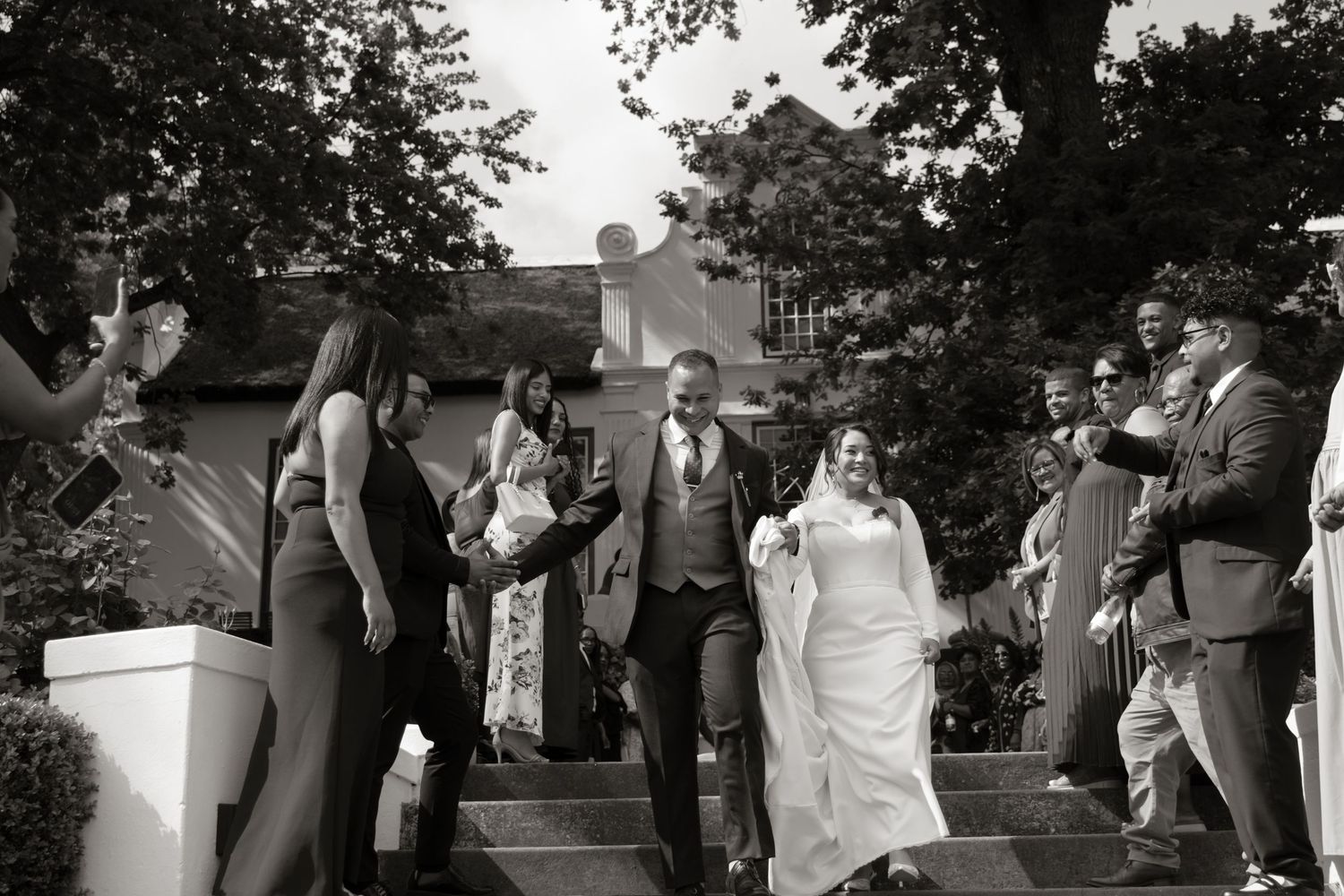 Black and white photo of a bride and groom walking down steps, surrounded by cheering wedding guests outdoors.