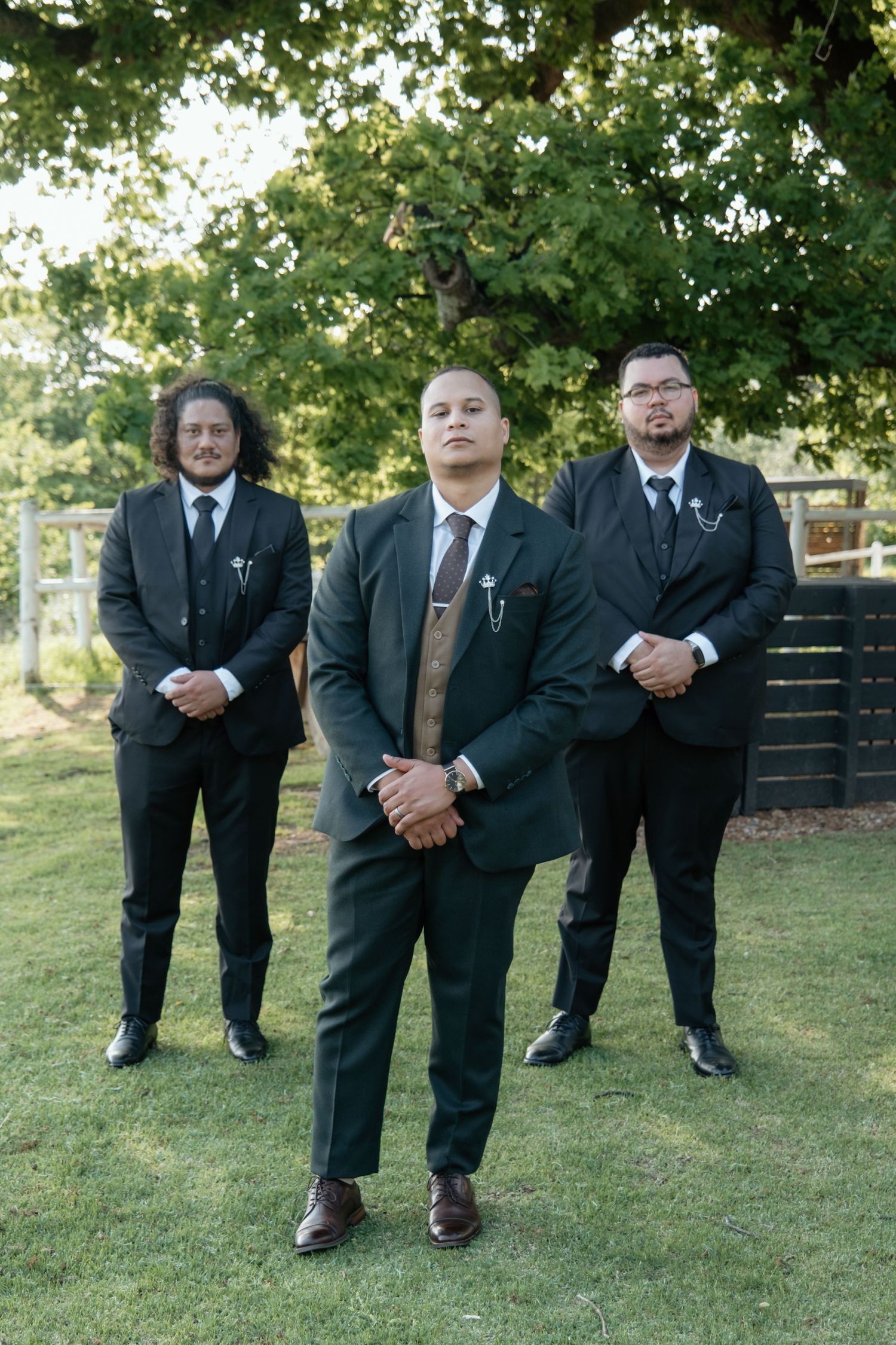 Three groomsmen in black suits posing outdoors under a tree at a wedding.