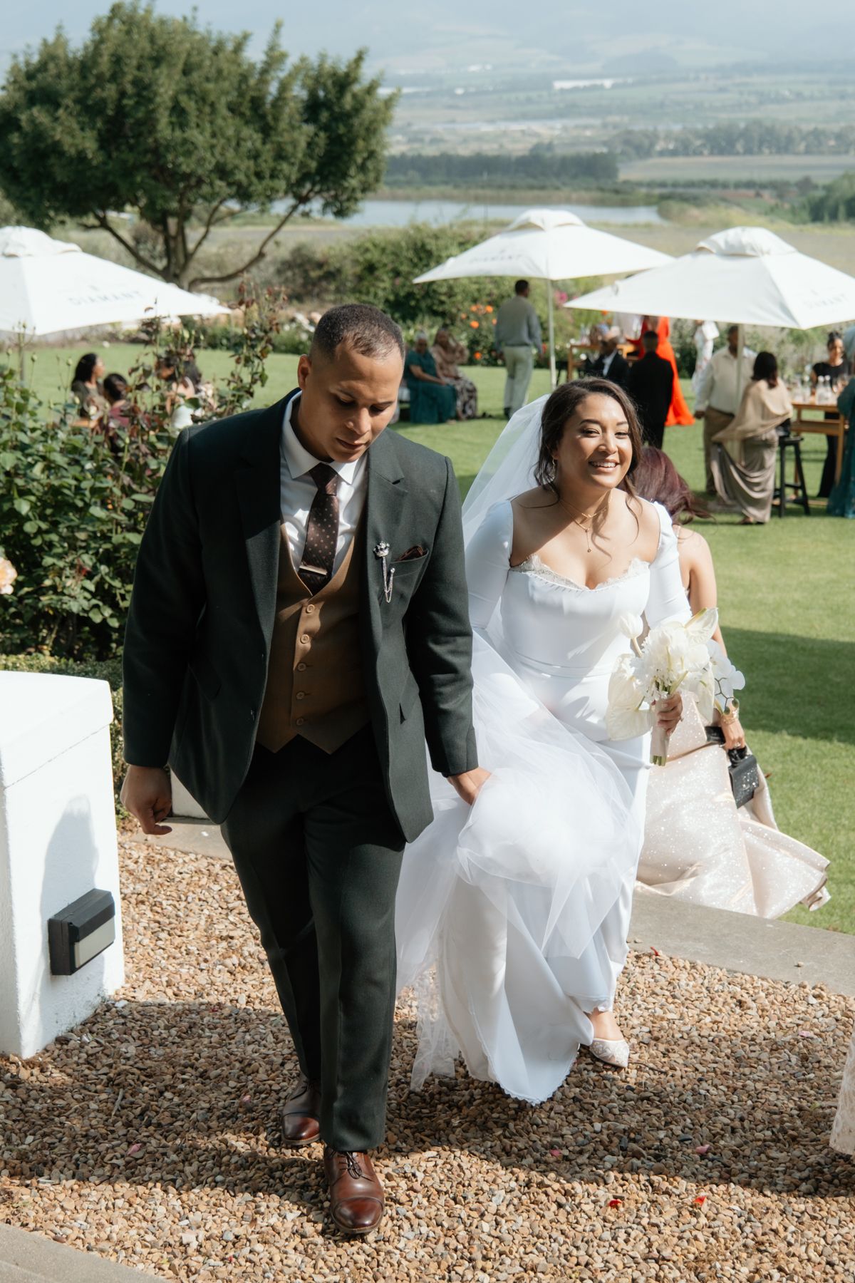 Smiling bride in white gown and groom in dark suit walk together at outdoor wedding venue with scenic views.