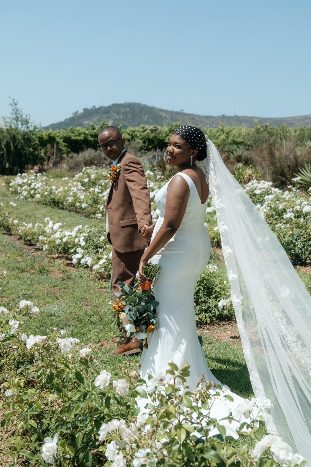 Black bride and groom holding hands in a white rose garden, bride's veil flowing, groom in brown suit, mountain backdrop.