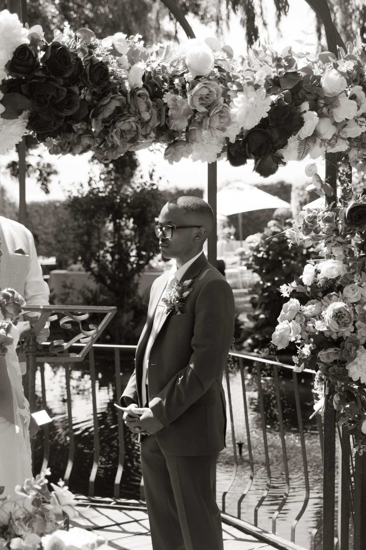 Groom in black suit stands under floral arch at outdoor wedding ceremony in black and white.