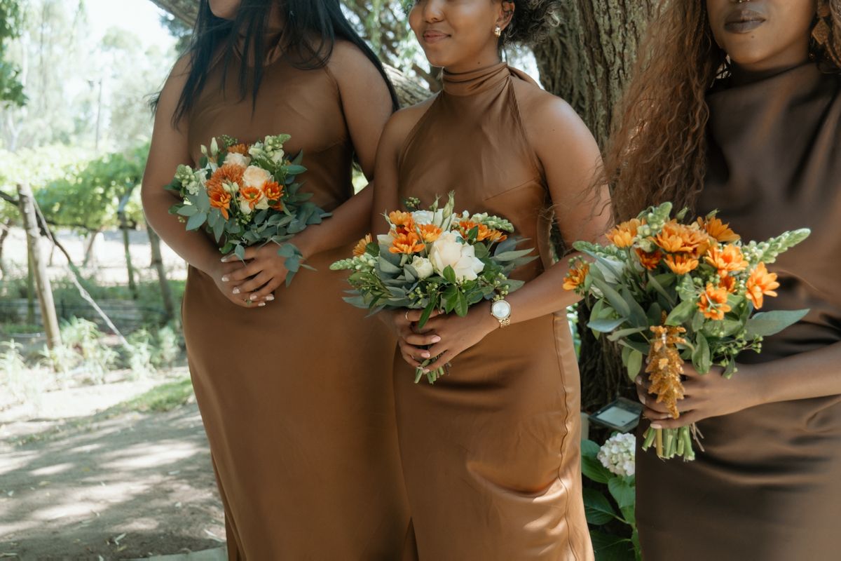 Three bridesmaids in brown satin dresses holding orange and white bouquets outdoors.