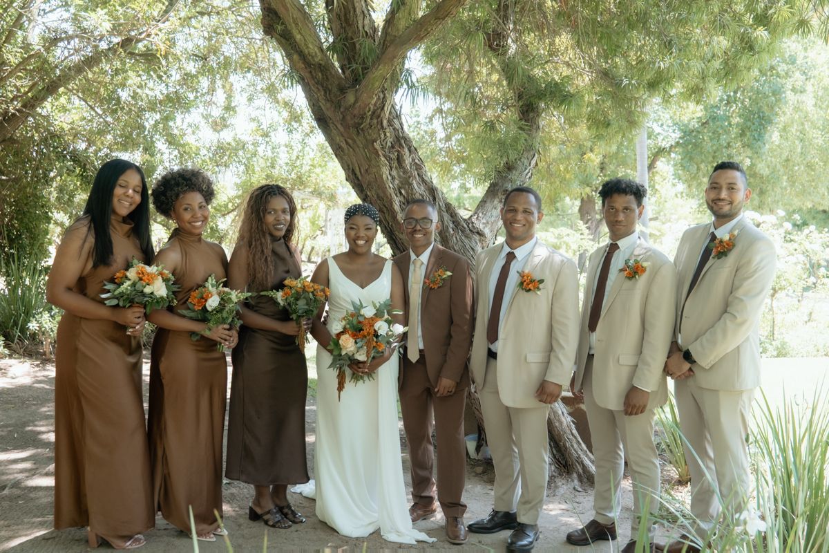 Wedding party poses under tree, bridesmaids in brown dresses, groomsmen in tan suits, bride in white gown.