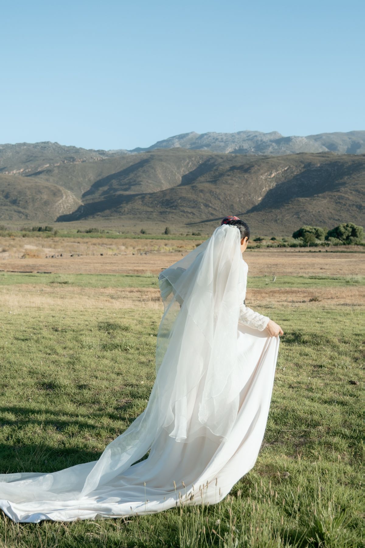 Bride in flowing white gown and veil stands in green meadow facing scenic mountain landscape.