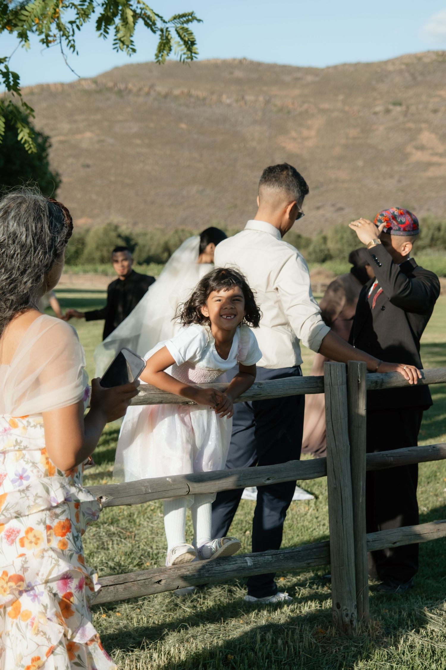 A young girl in white dress leans on wooden fence at outdoor wedding ceremony with mountains in background.