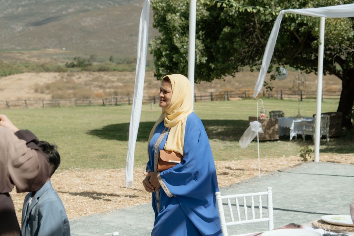 Woman in blue abaya and yellow hijab smiles at outdoor wedding reception with mountains behind.