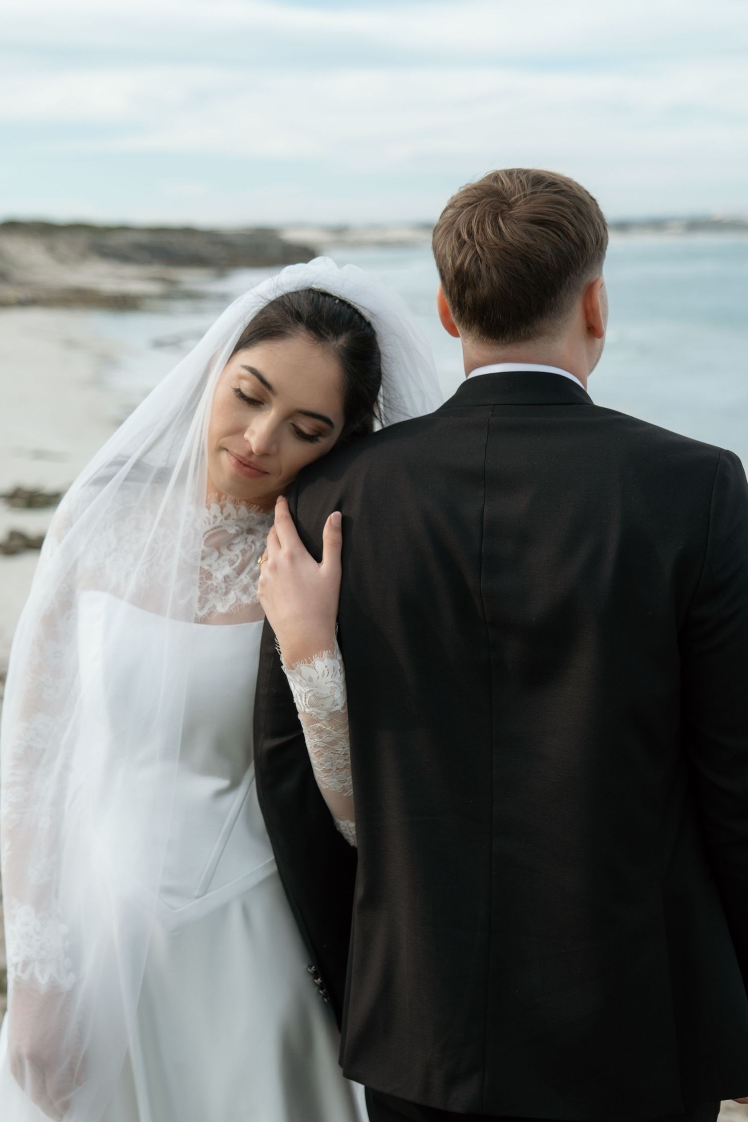 Bride in white lace gown and veil rests head on groom's shoulder near a sandy beach under cloudy skies.