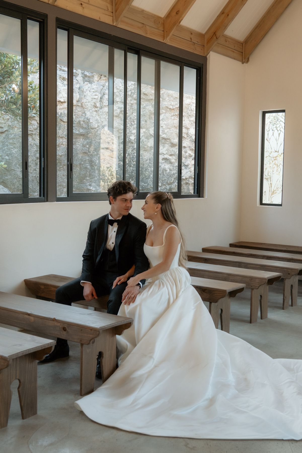 Bride and groom sitting close together on wooden bench in sunlit chapel with stone wall windows.