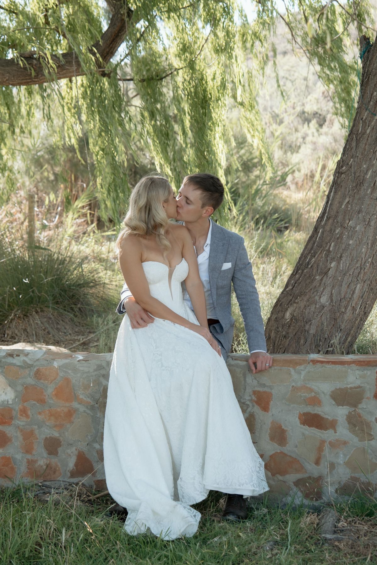 Newlyweds sharing a kiss beside a rustic stone wall under a willow tree outdoors.