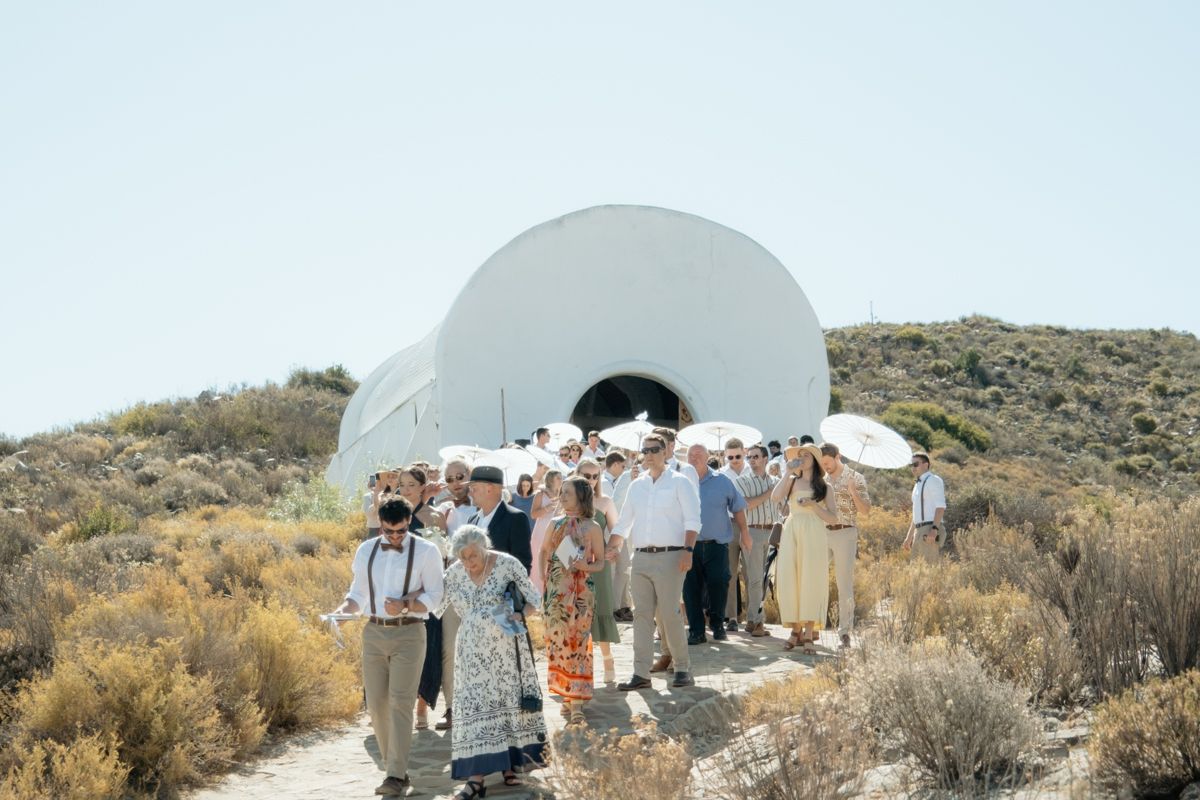 Wedding guests gather outside a white dome-shaped chapel on a sunny hillside ceremony.