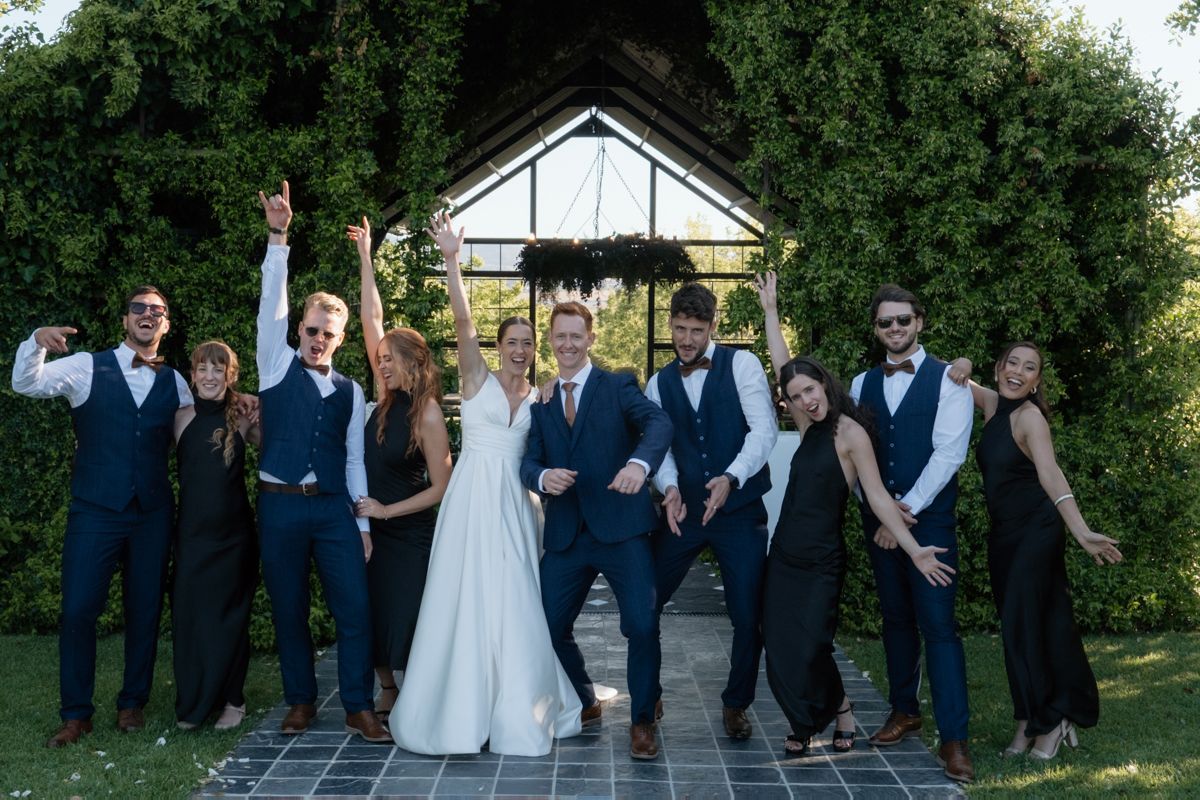Wedding party in navy suits and black dresses celebrating joyfully in front of a green ivy-covered arch.