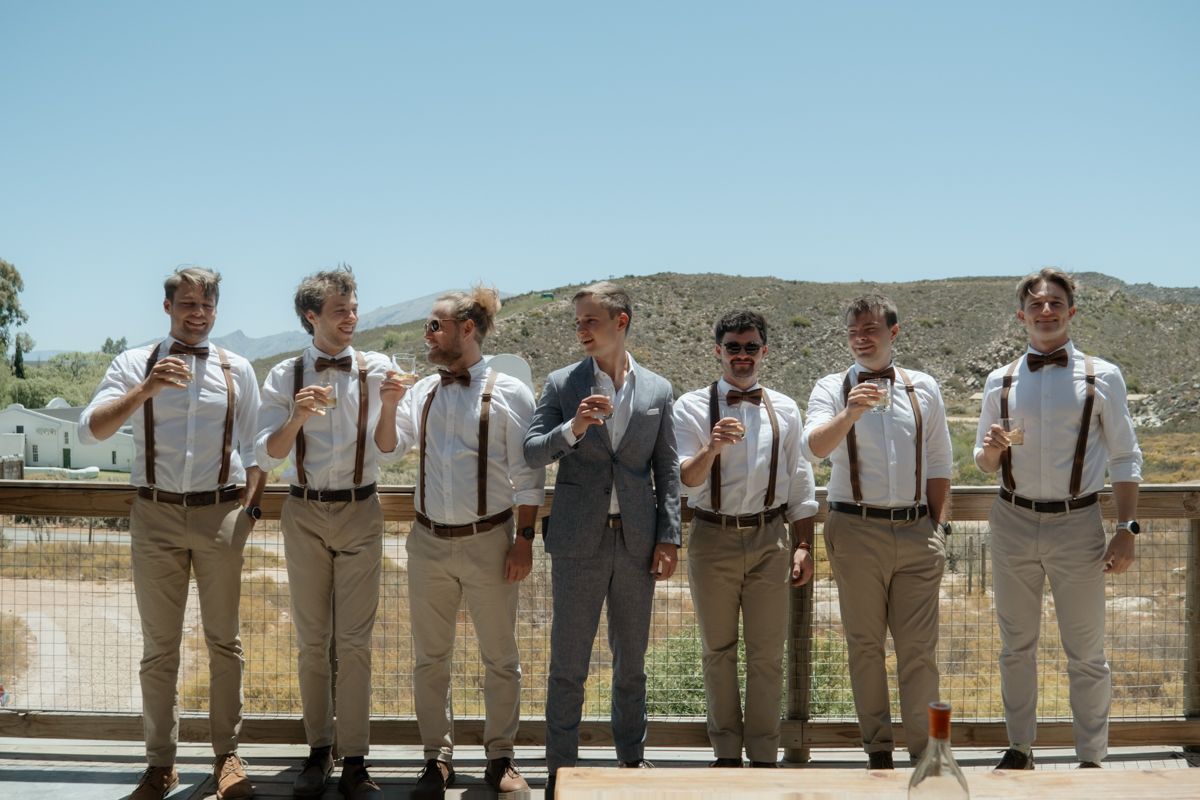 Groom and groomsmen in tan pants, suspenders, and bow ties posing outdoors at a sunny wedding.