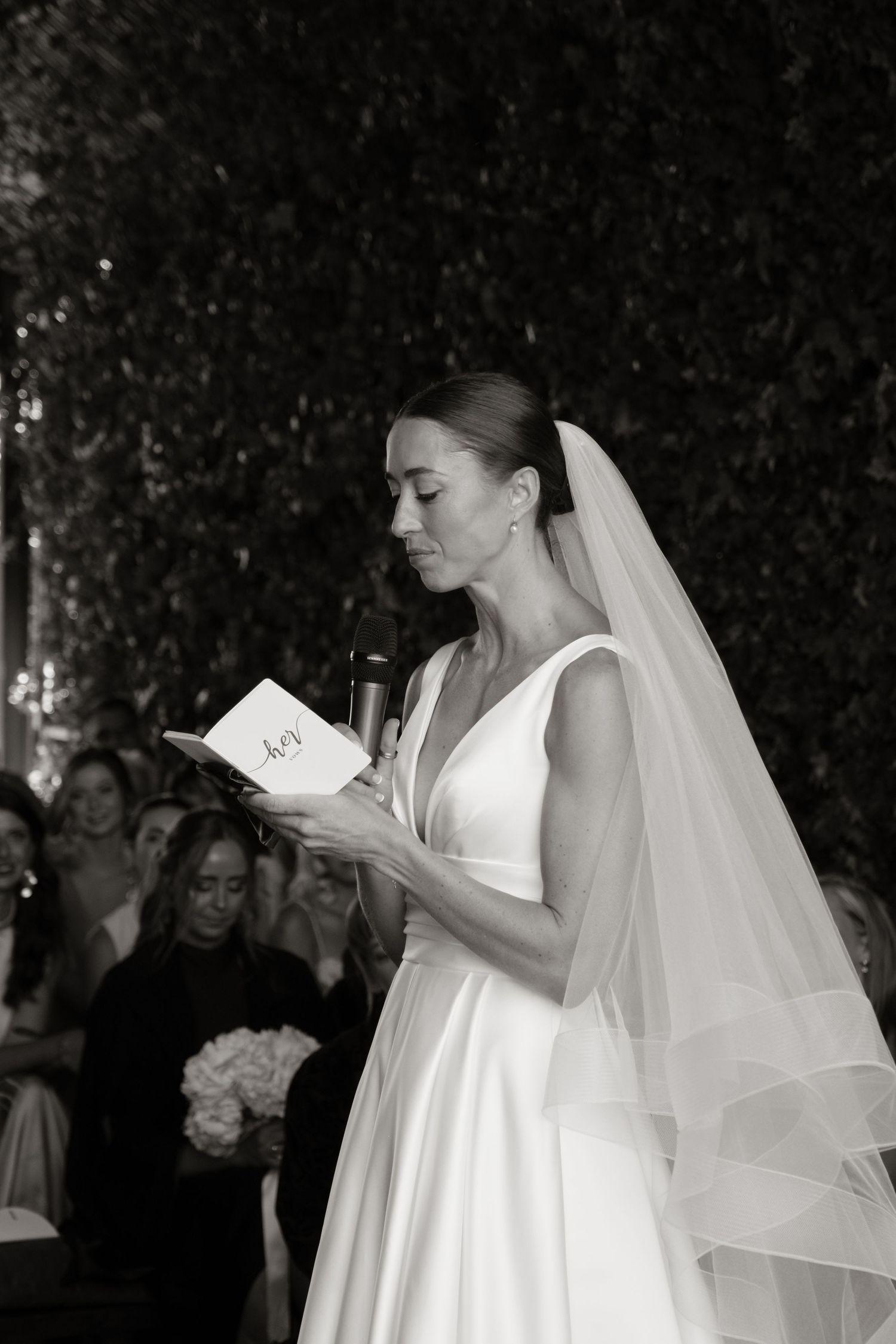 Bride in white gown and veil reading vows from booklet at wedding ceremony.