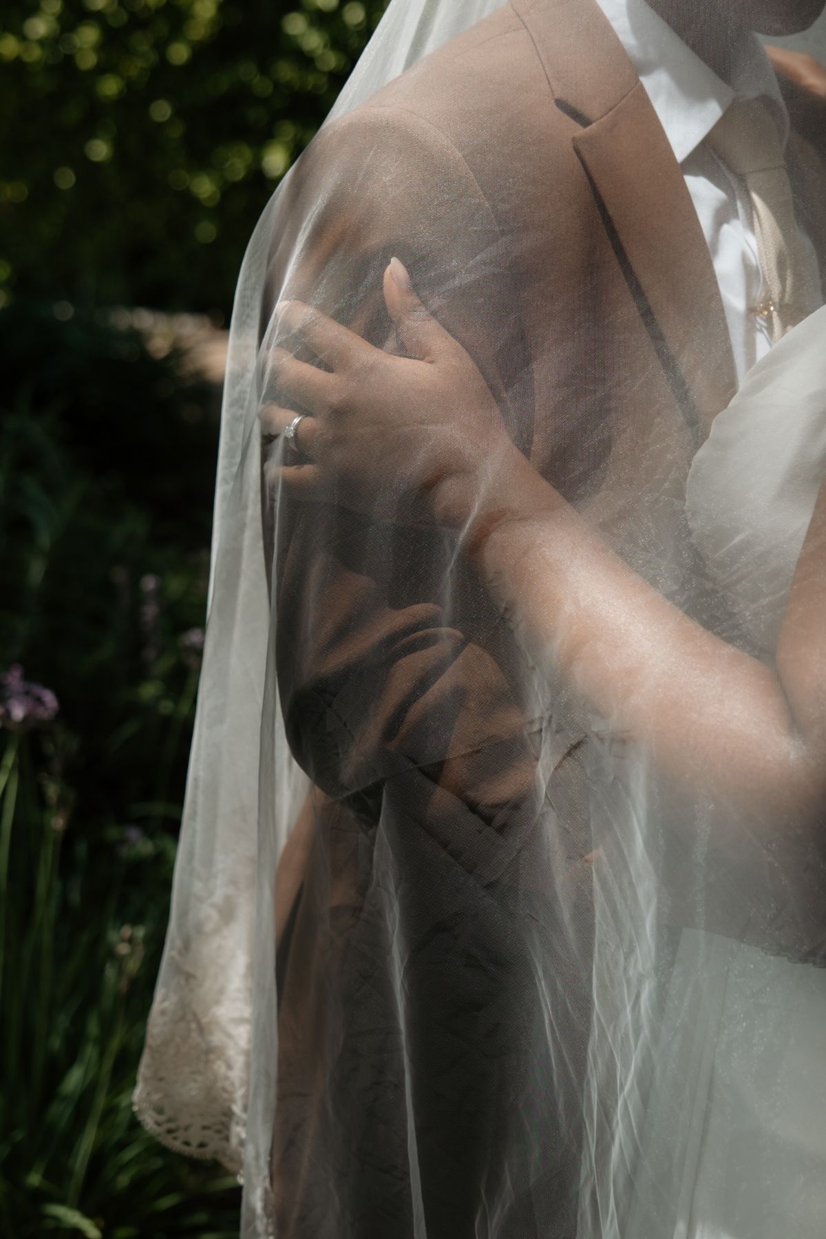 Close-up of bride's hand with wedding ring gently holding groom's arm through a delicate lace veil.