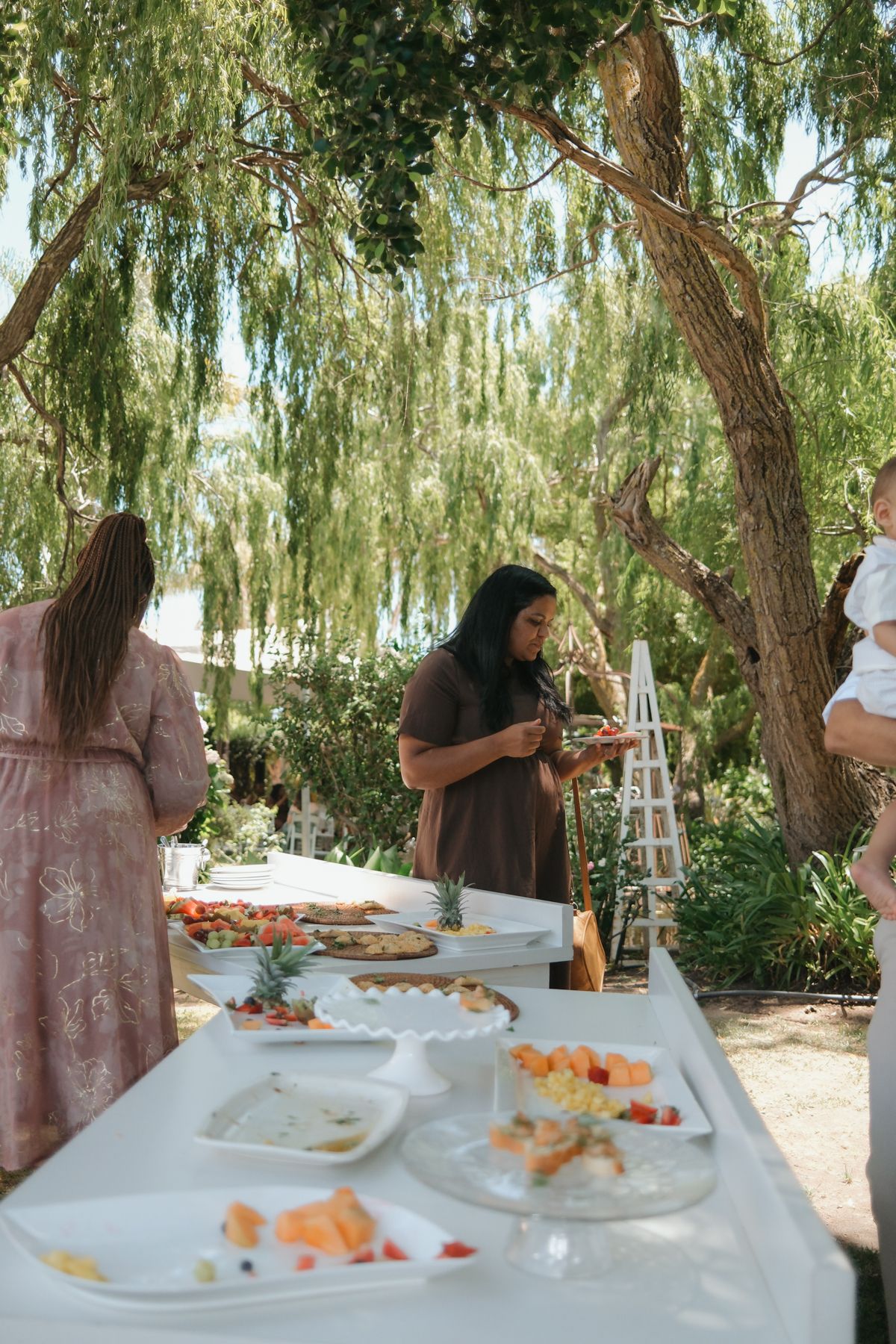 Guests serving themselves at an elegant outdoor wedding buffet table with fresh fruit and appetizers under willow trees.