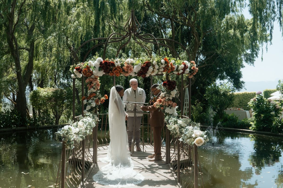 Bride in white gown walks toward floral arch over water at elegant outdoor wedding ceremony.